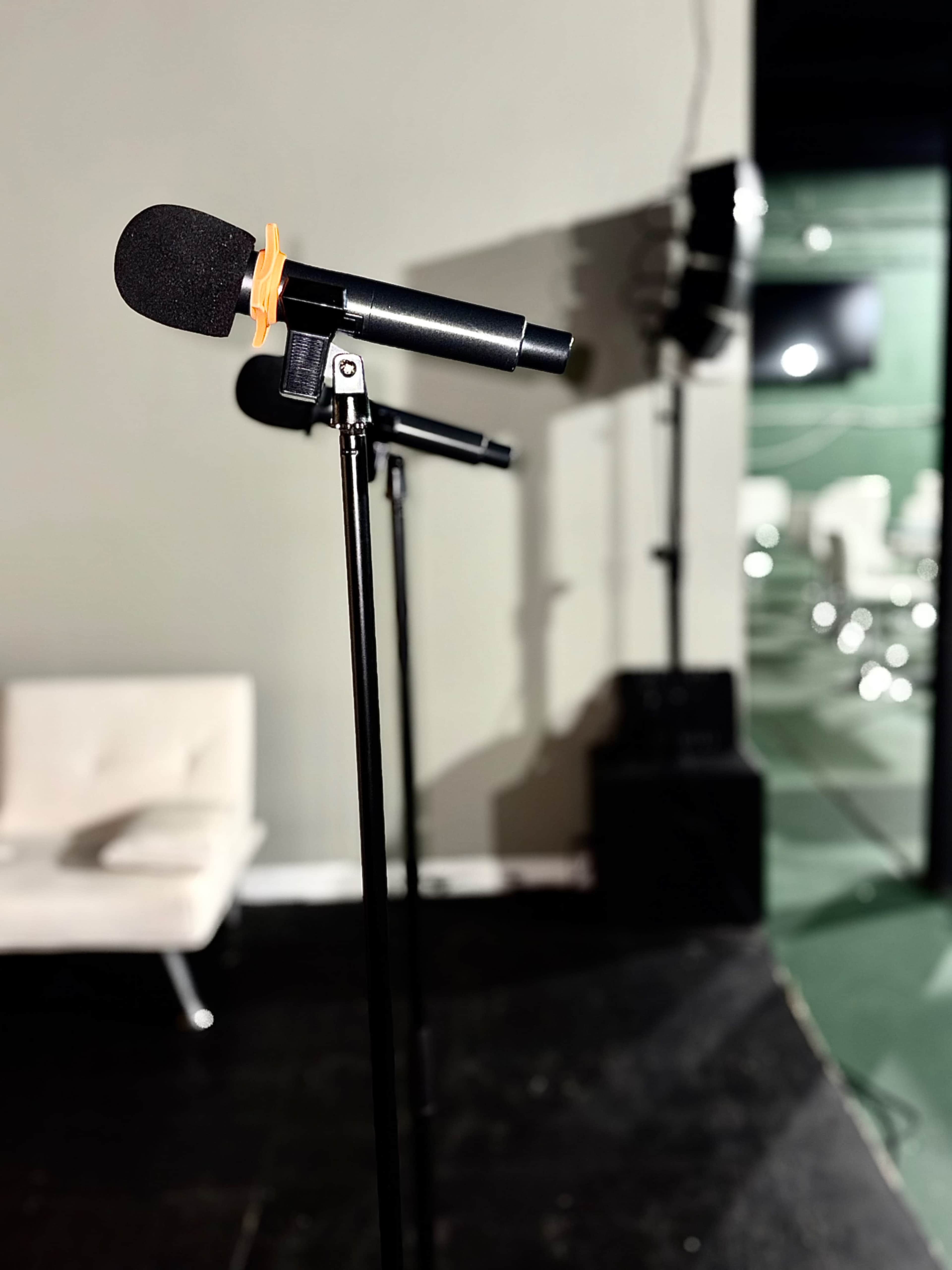 A microphone stands on a black stand in a partially empty room with chairs in the background.