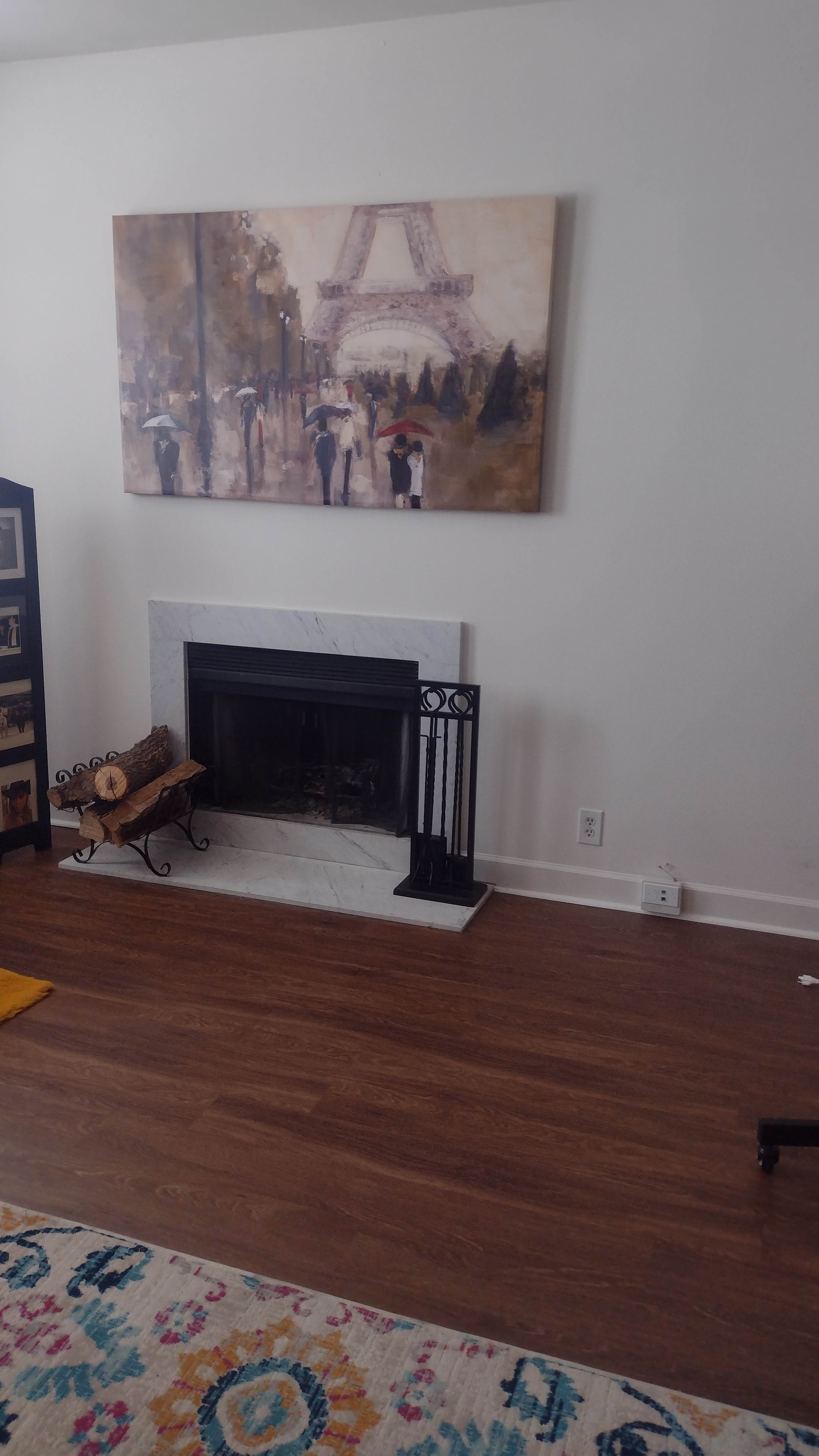 The image shows a living room with a white fireplace, a black decorative screen, a painting of the Eiffel Tower on the wall, and a log holder beside the fireplace.