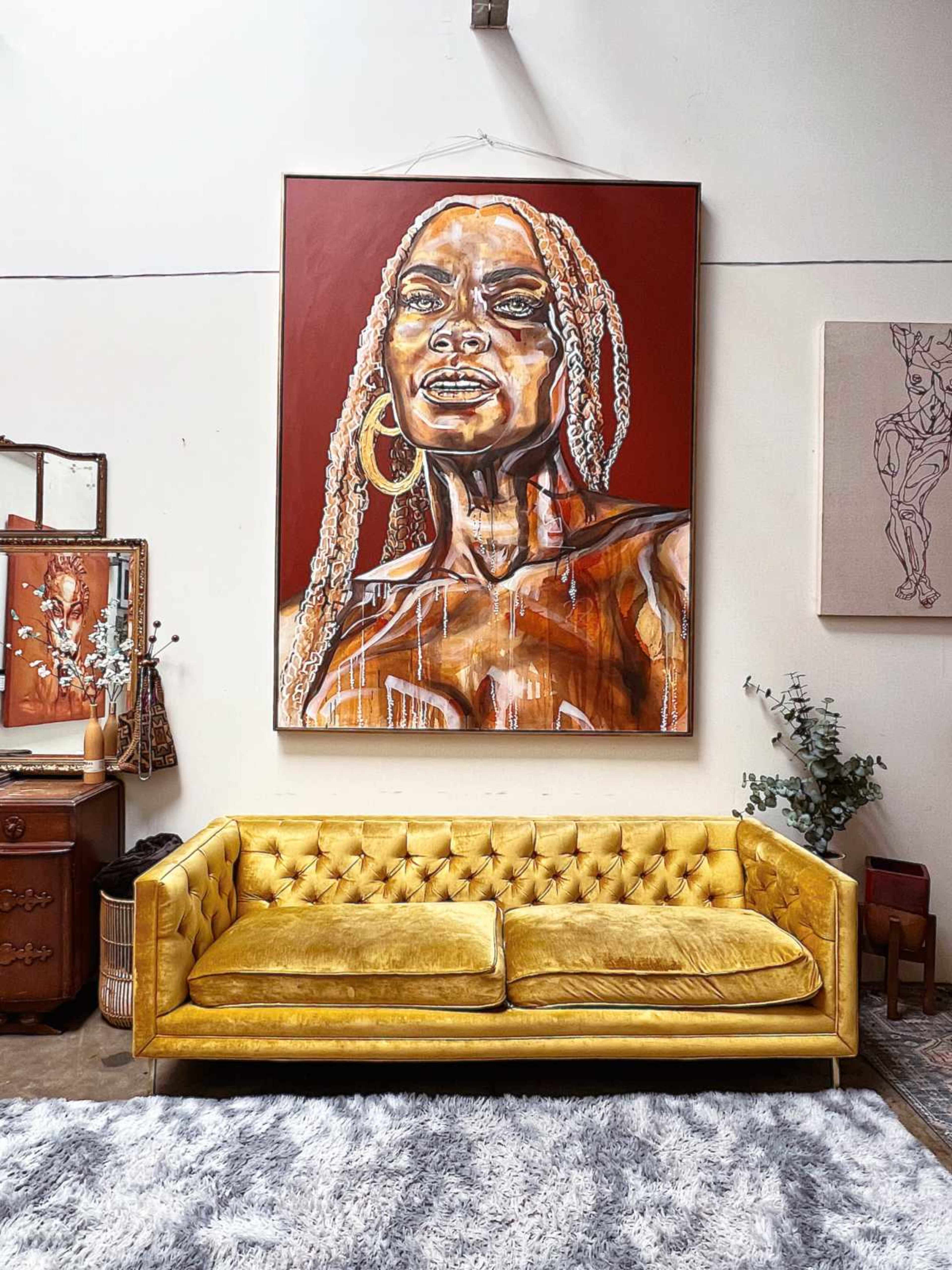 A yellow velvet couch sits in front of a large artwork featuring a portrait of a woman, displayed on a light-colored wall.