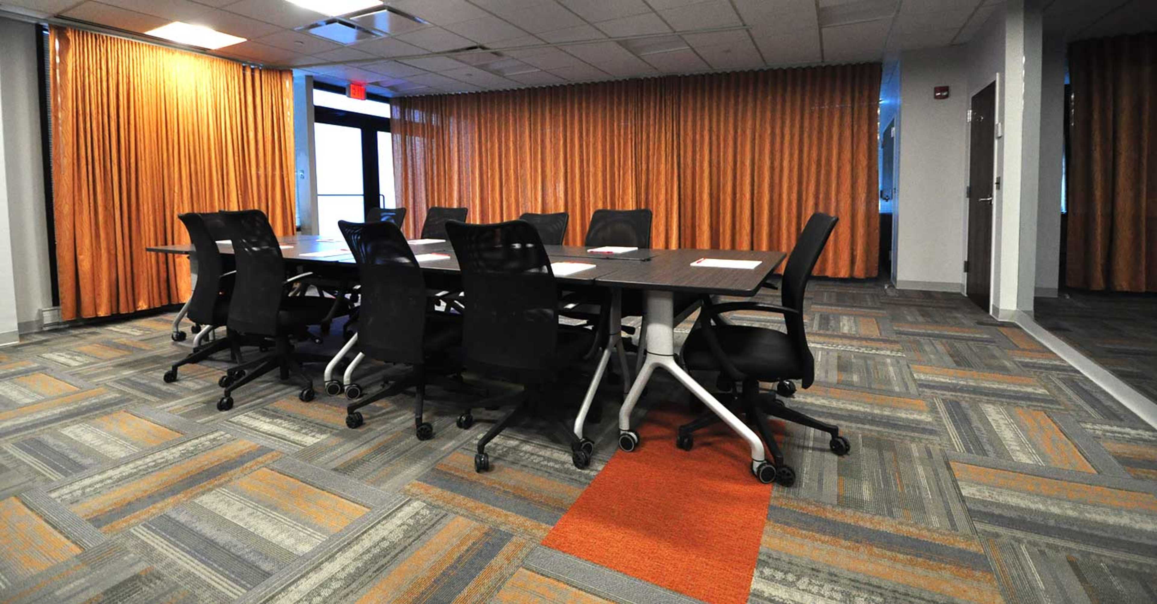 A conference room features a large table surrounded by multiple black chairs, with orange curtains covering the windows.
