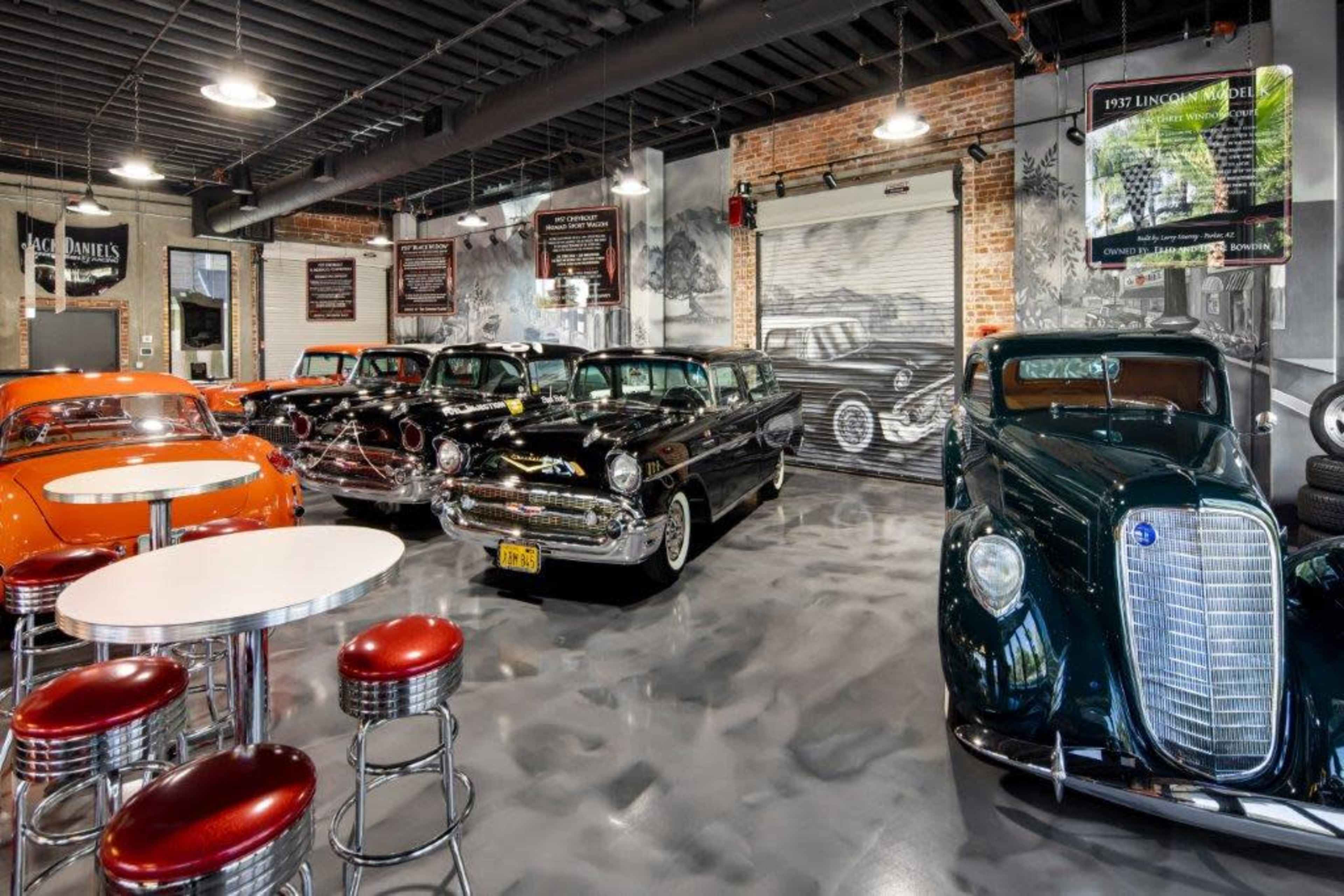A vintage car showroom featuring several classic cars, including two black sedans, an orange vehicle, and a green model, set against a backdrop of brick walls and car-themed decor.