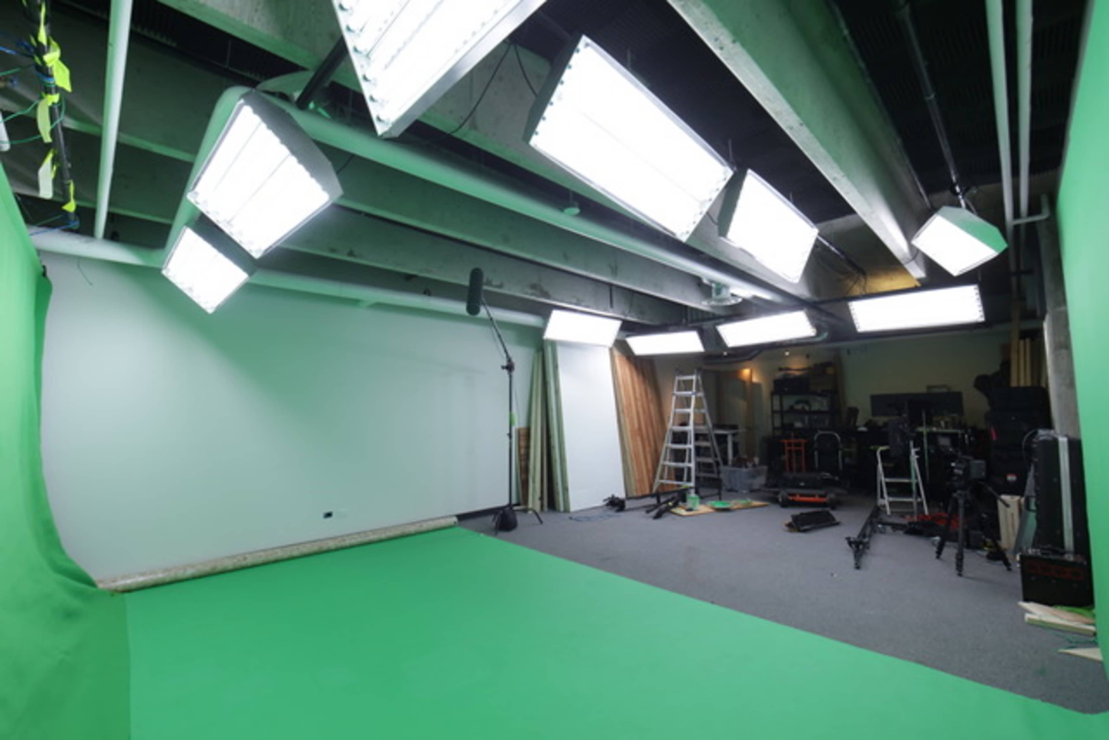 A green screen studio is equipped with bright lights, a ladder, and various camera equipment.