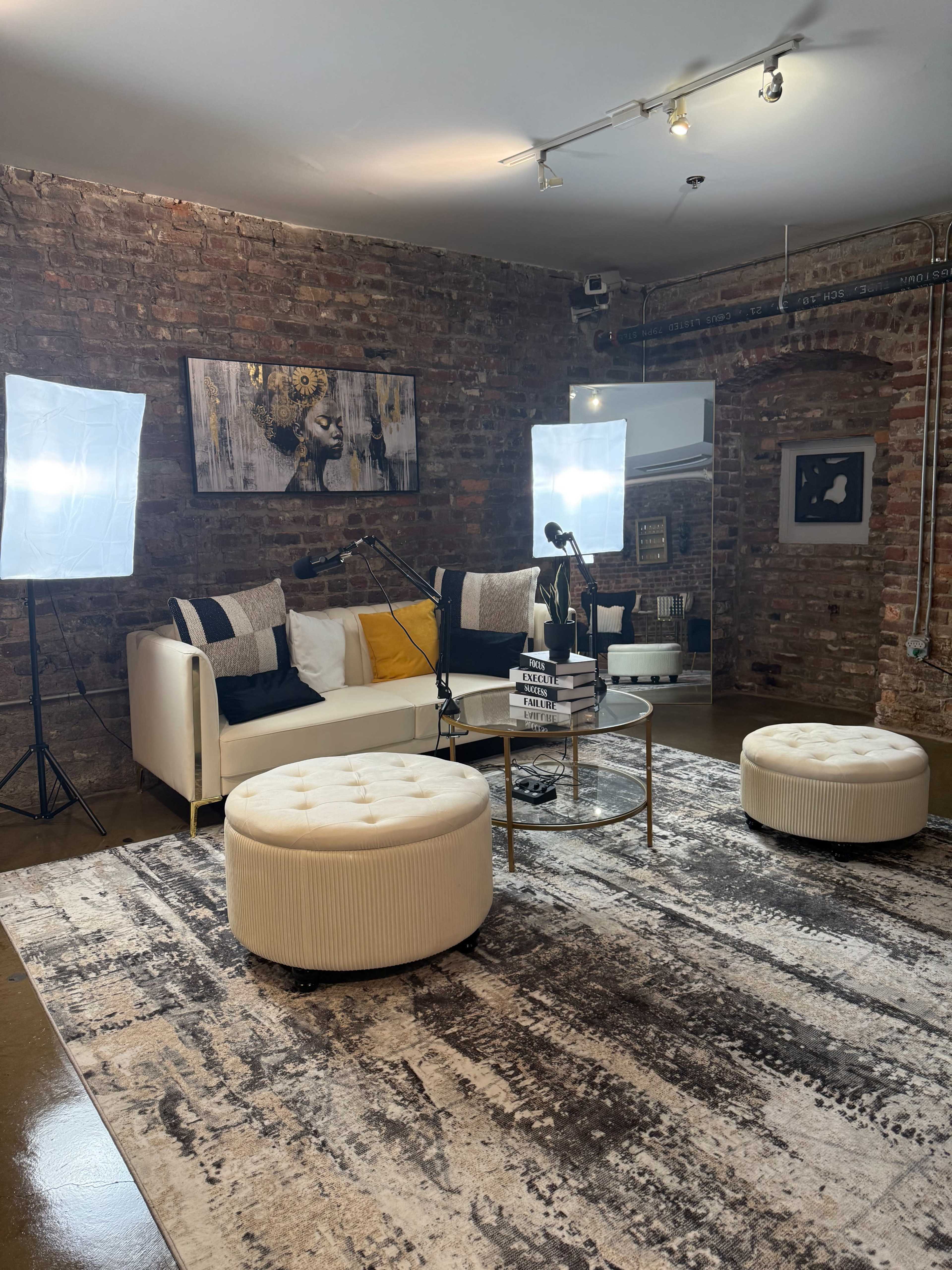 The image shows a modern living room setting with a white couch, two round ottomans, a coffee table, and studio lighting in a brick-walled environment.