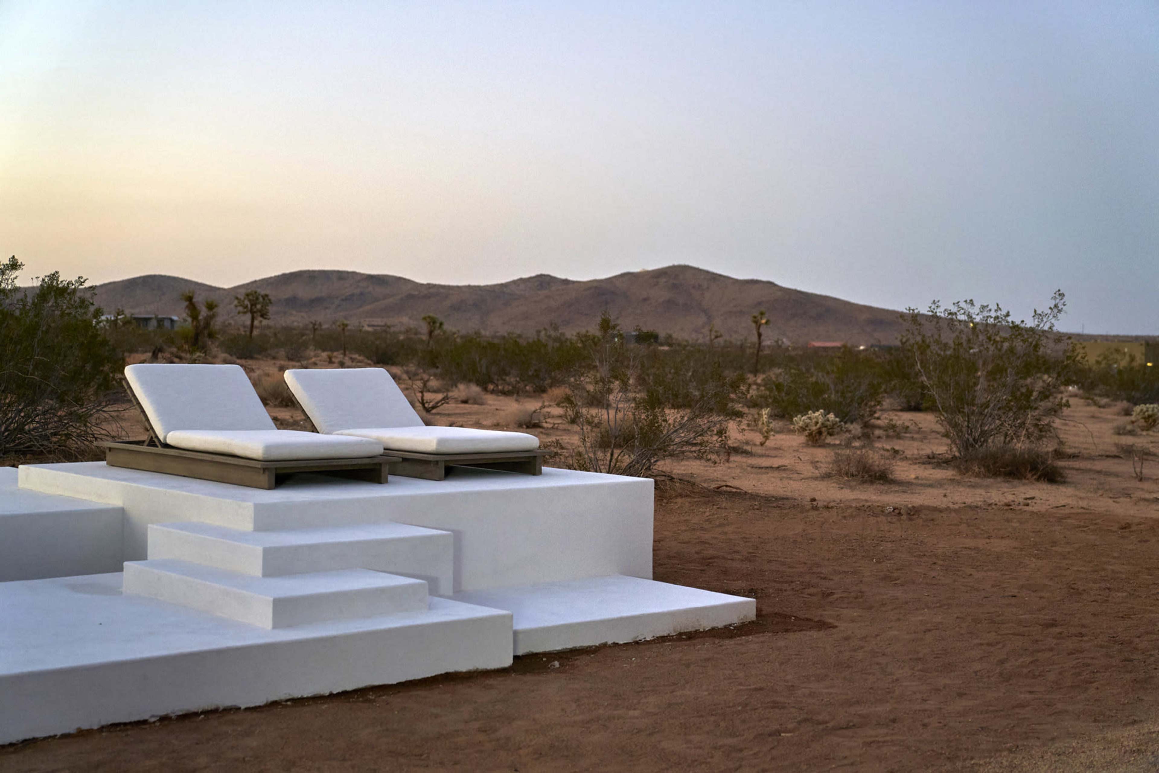 Two white lounge chairs are positioned on a raised platform in a barren desert landscape with rolling hills in the background.