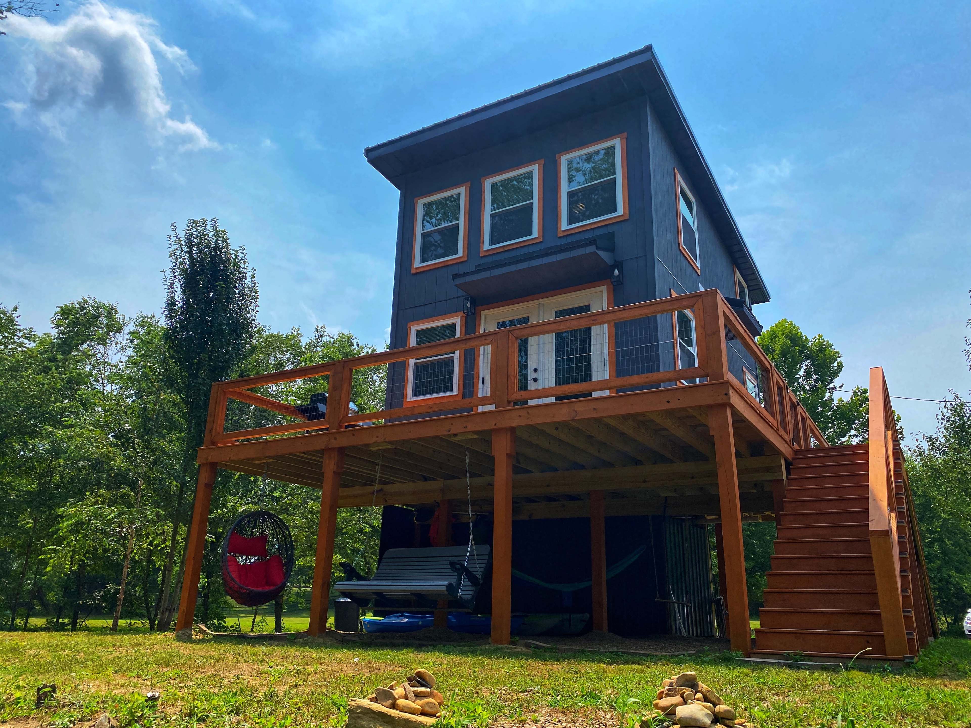 A modern treehouse with a wooden deck and stairs stands elevated among green trees on a sunny day.