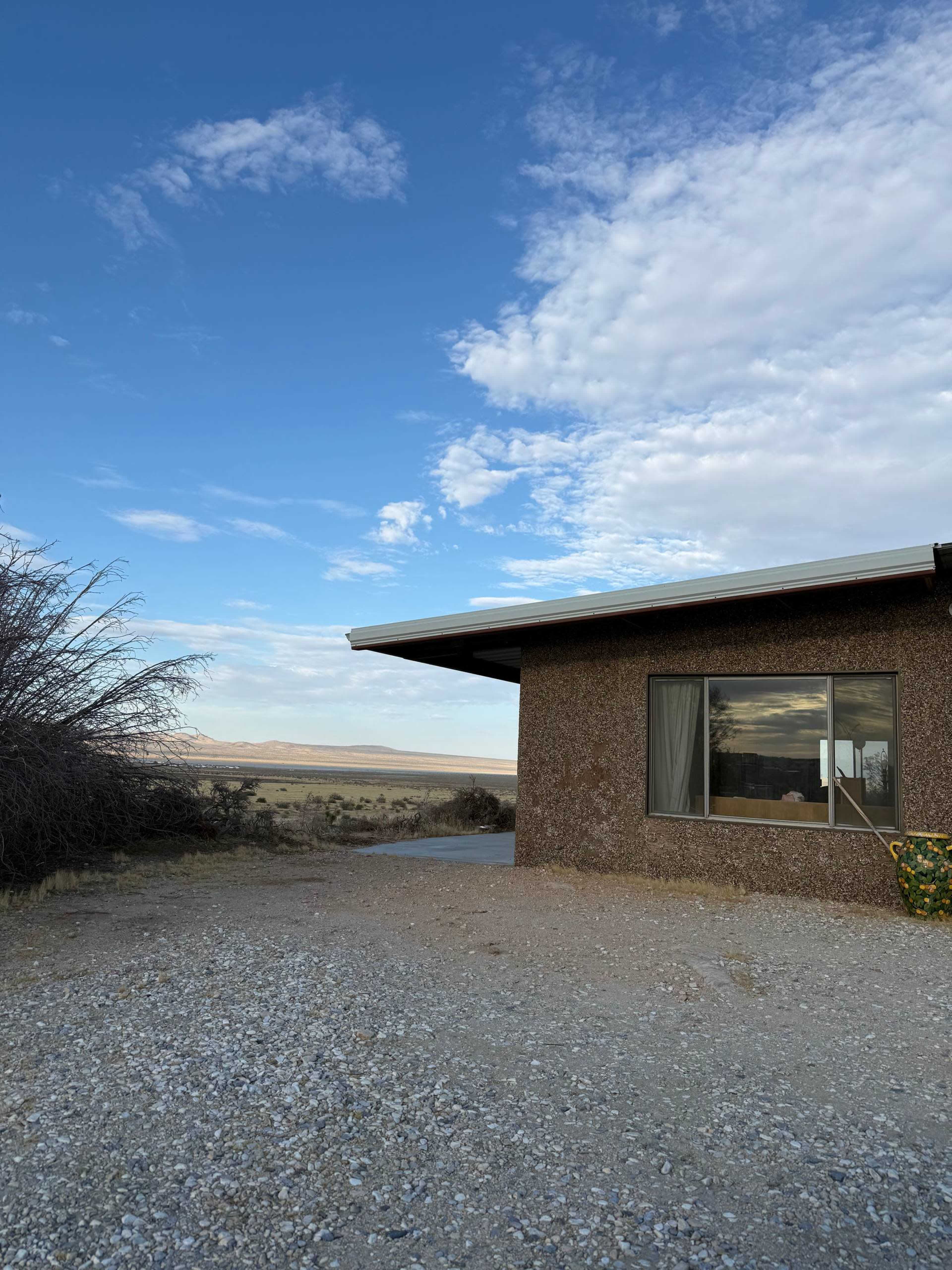 A single-story building with a textured exterior and a large window is situated in a gravel area, overlooking a vast open landscape under a partly cloudy sky.
