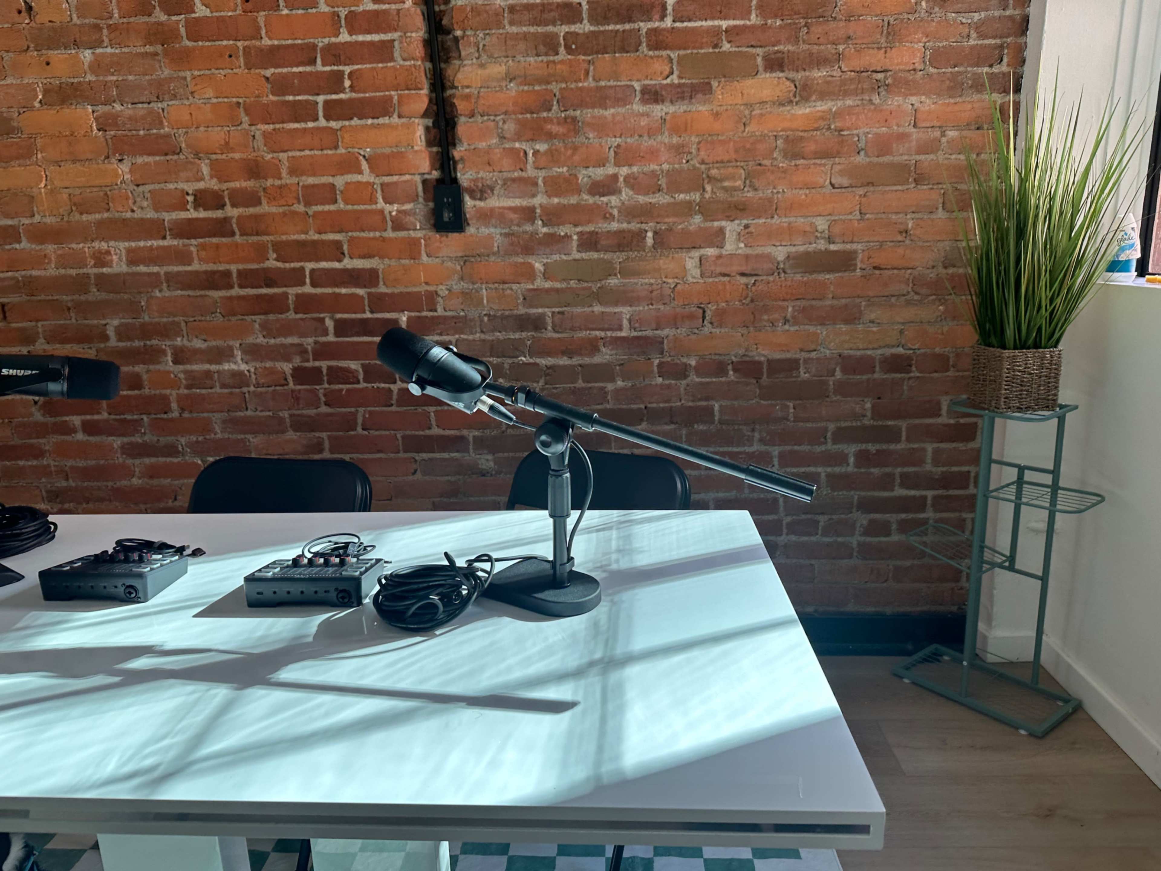 A table with microphones and audio equipment is set against a wall of exposed brick with a potted plant in the corner.