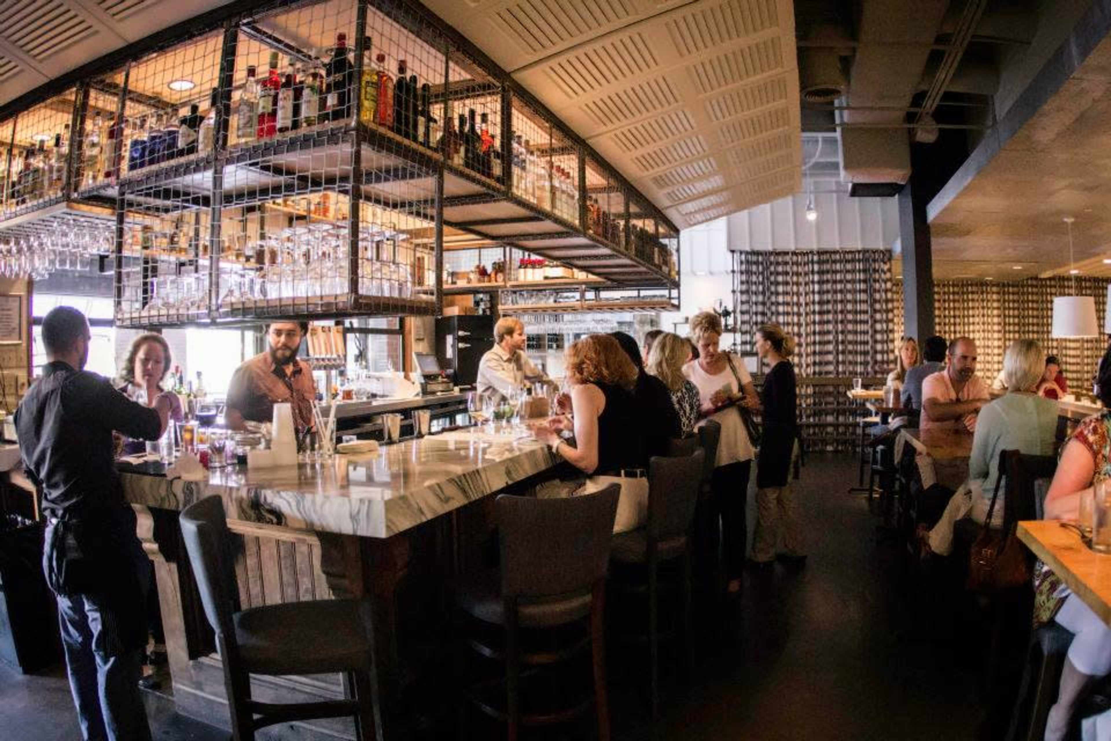 A bustling bar area in a restaurant features patrons seated at high-top tables and the bar, with shelves displaying various liquor bottles and glasses overhead.