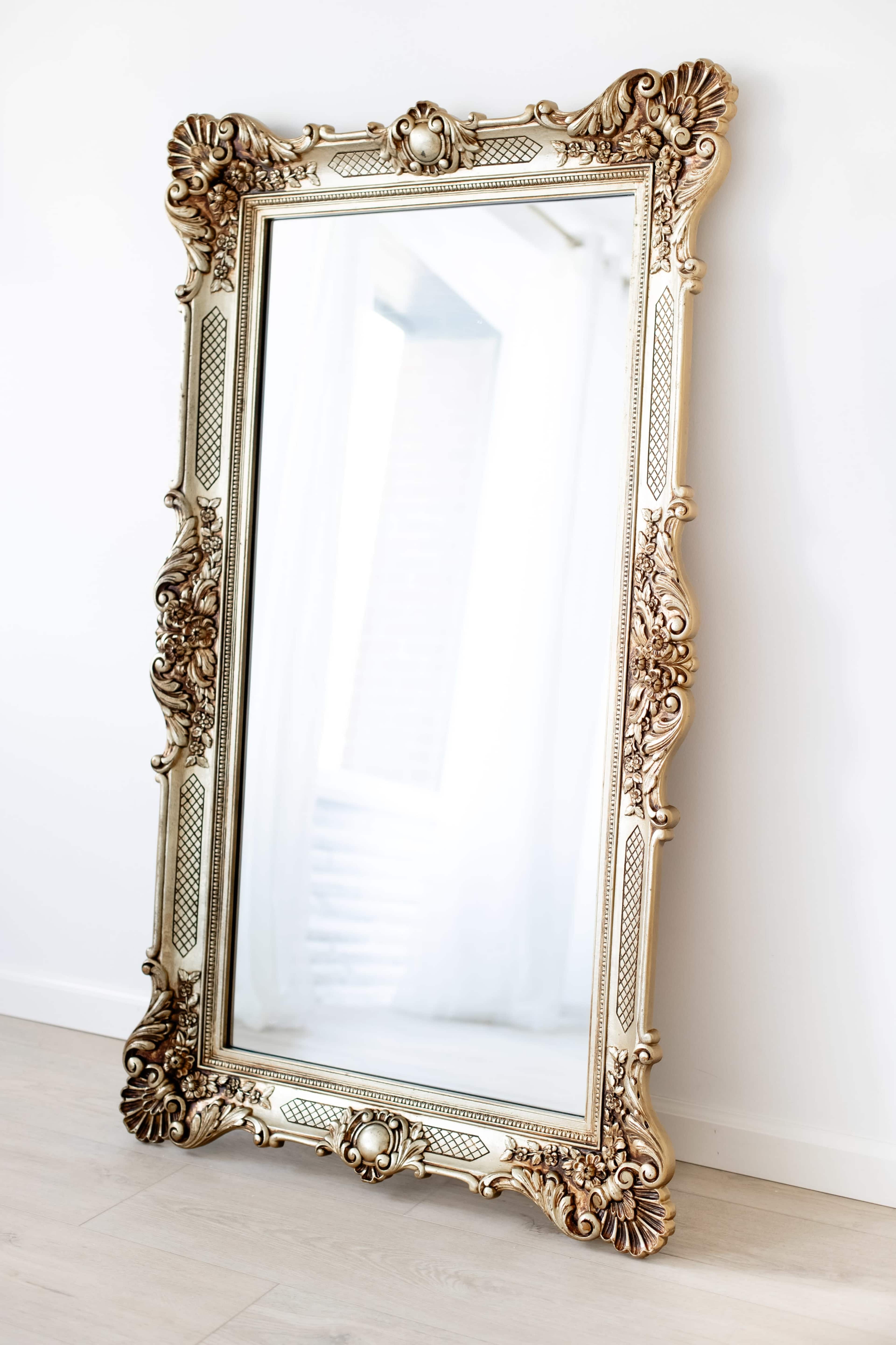 A large, ornate mirror with a decorative gold frame leans against a white wall on a light wooden floor.