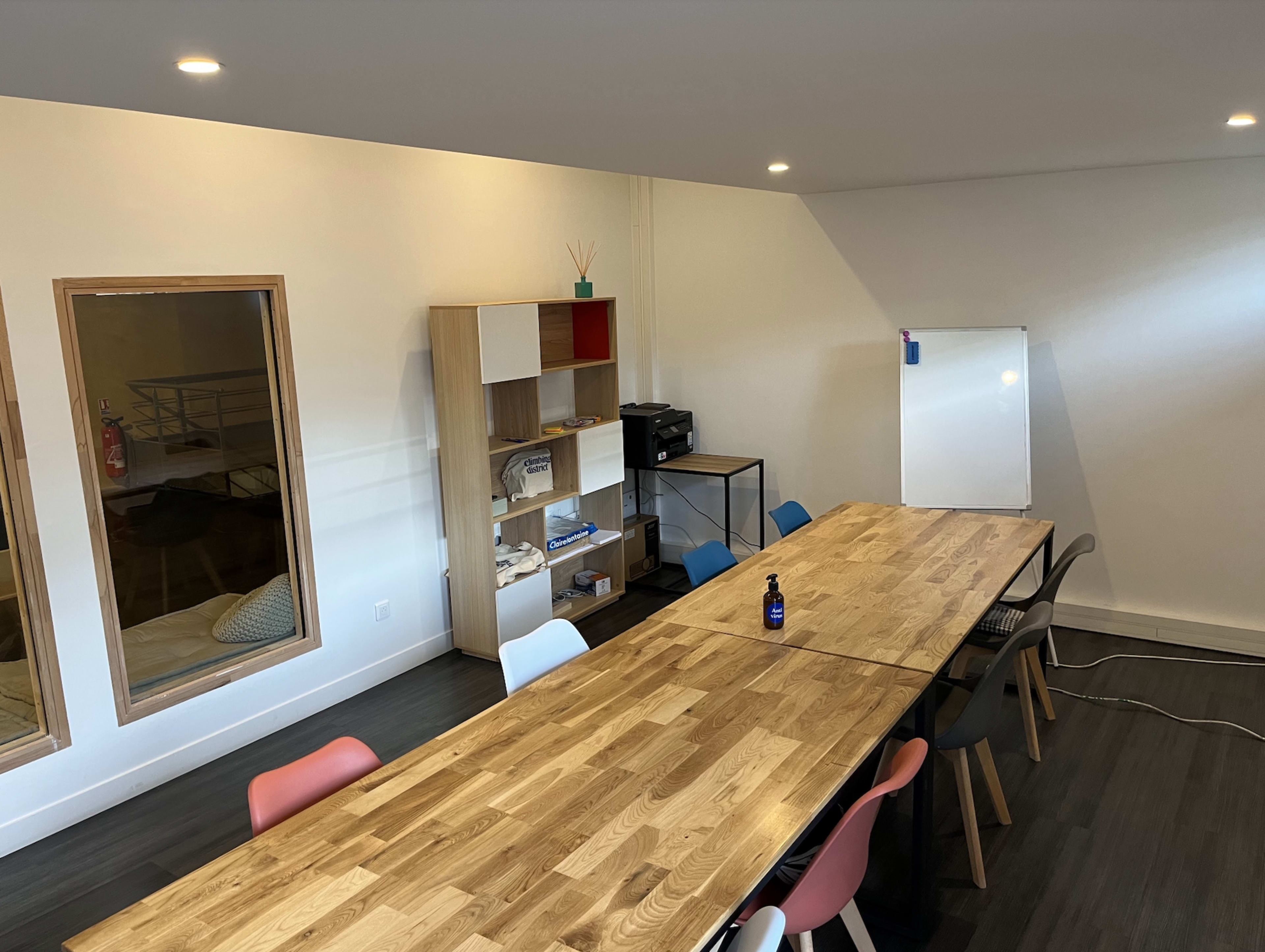 A modern meeting room features a long wooden table surrounded by colorful chairs, a whiteboard, and a shelving unit with various items.