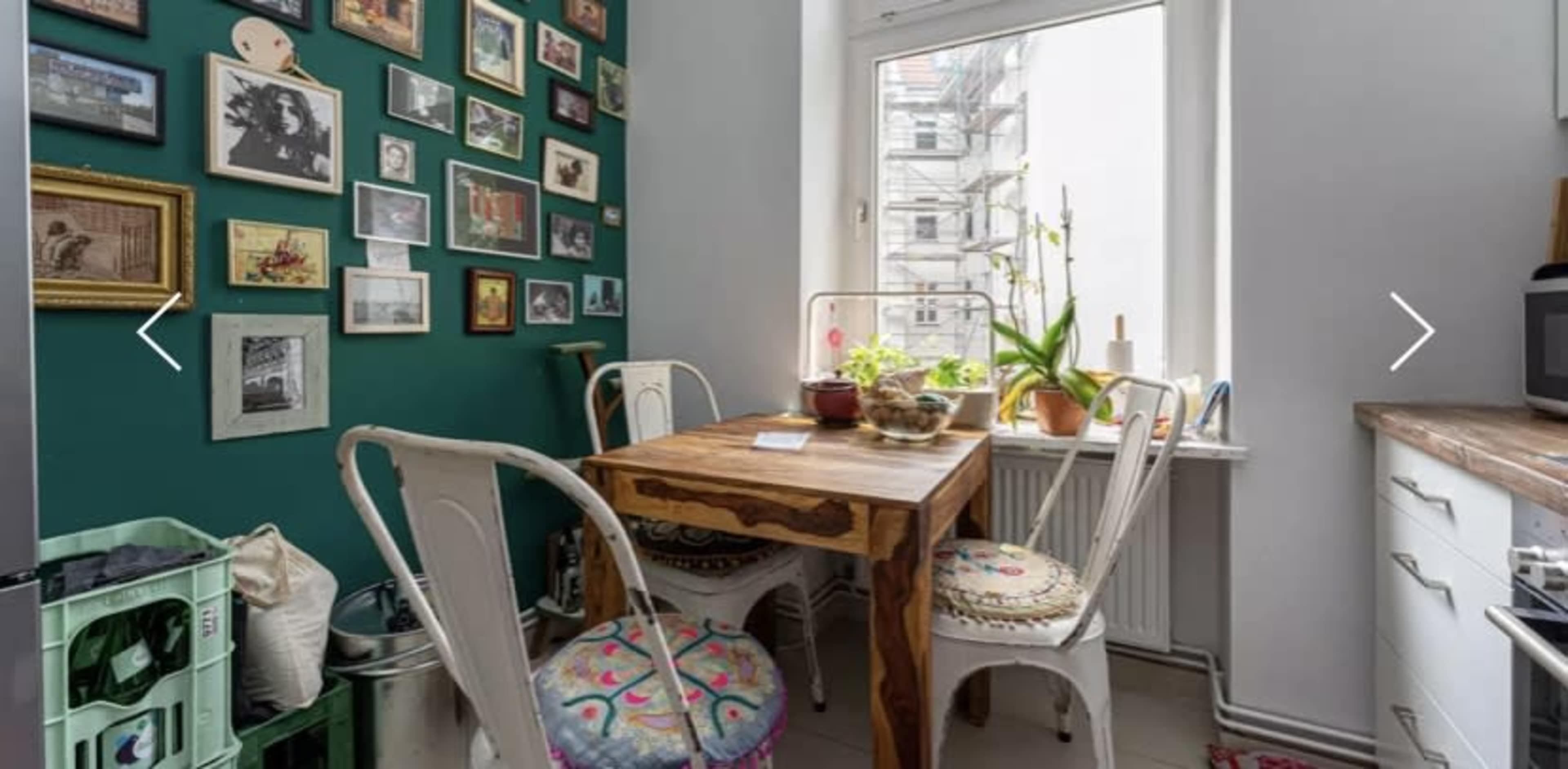 A small kitchen with a wooden table and four chairs, a green wall adorned with framed pictures, and a window with potted plants.