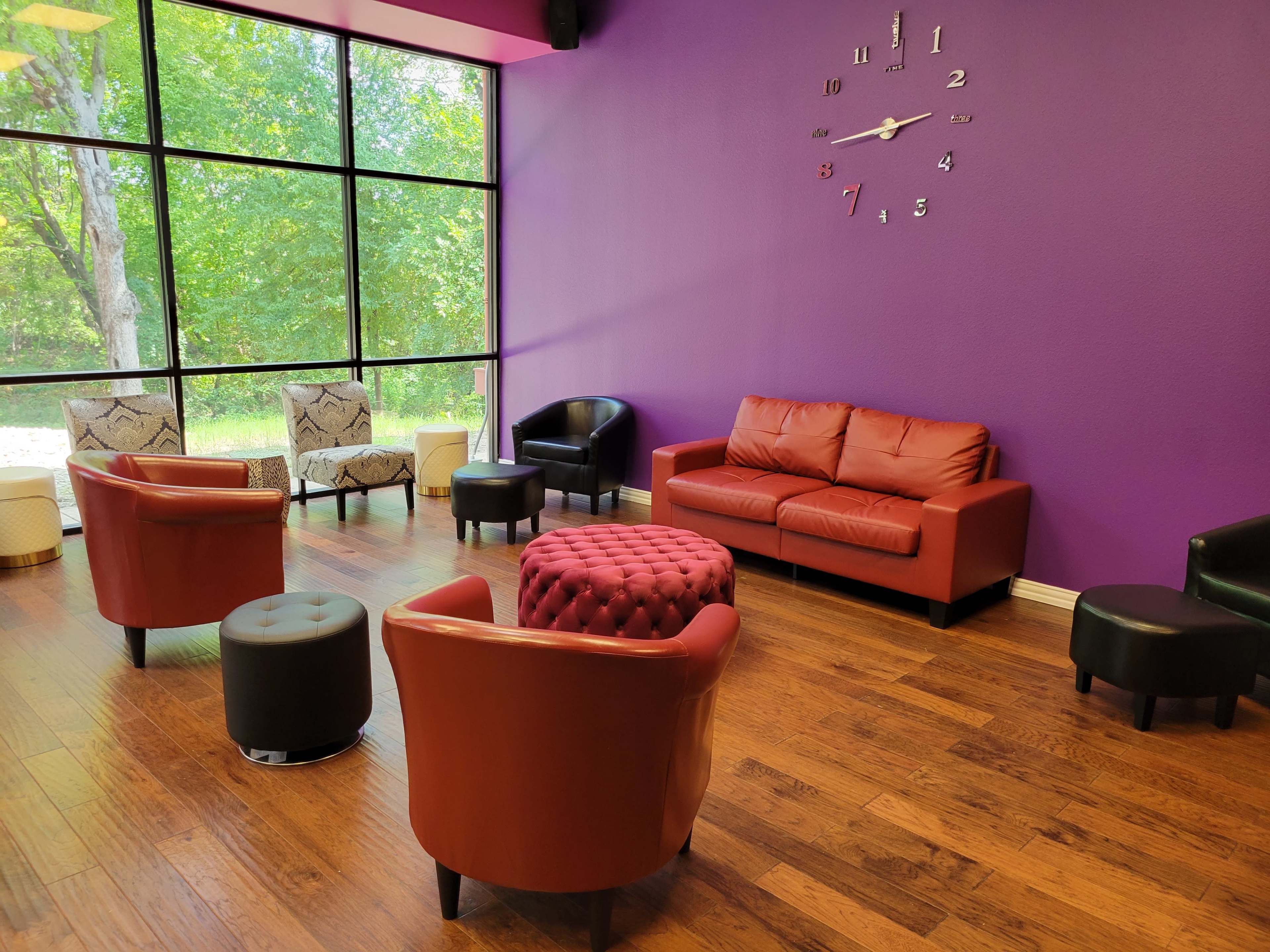 A seating area features red and black chairs, a brown couch, and a round ottoman, set against a purple wall and large windows overlooking greenery.