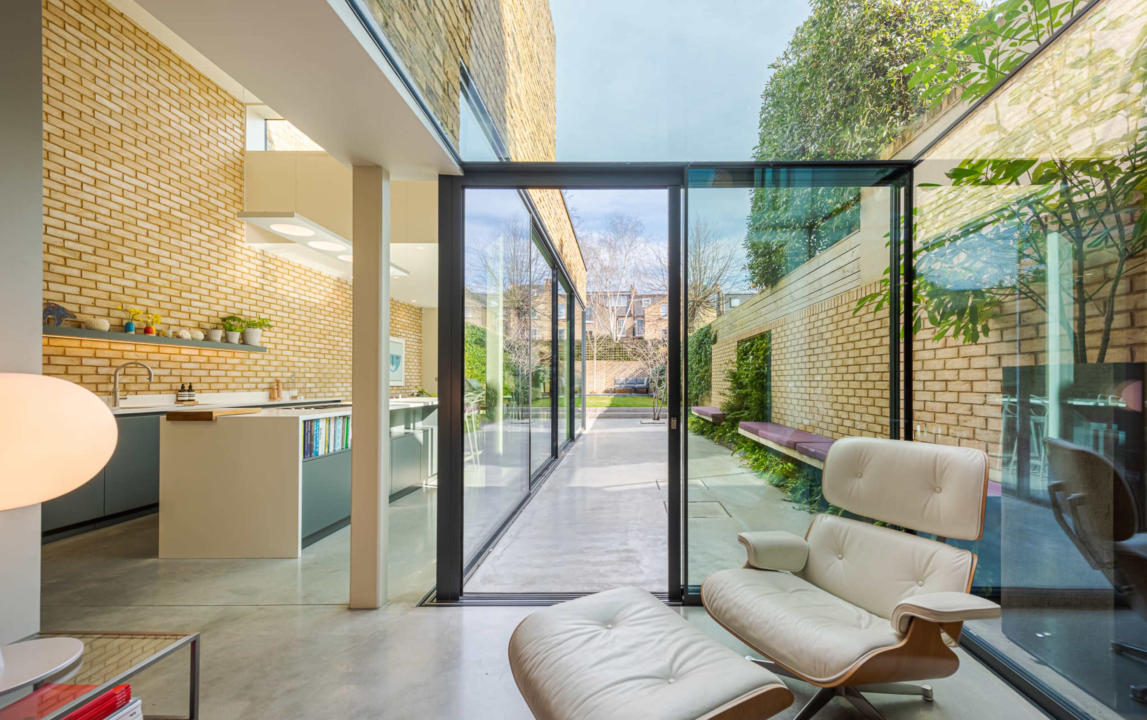 A modern interior featuring a glass-walled living space that opens to a landscaped garden, showcasing a minimalist design with a kitchen and seating area.