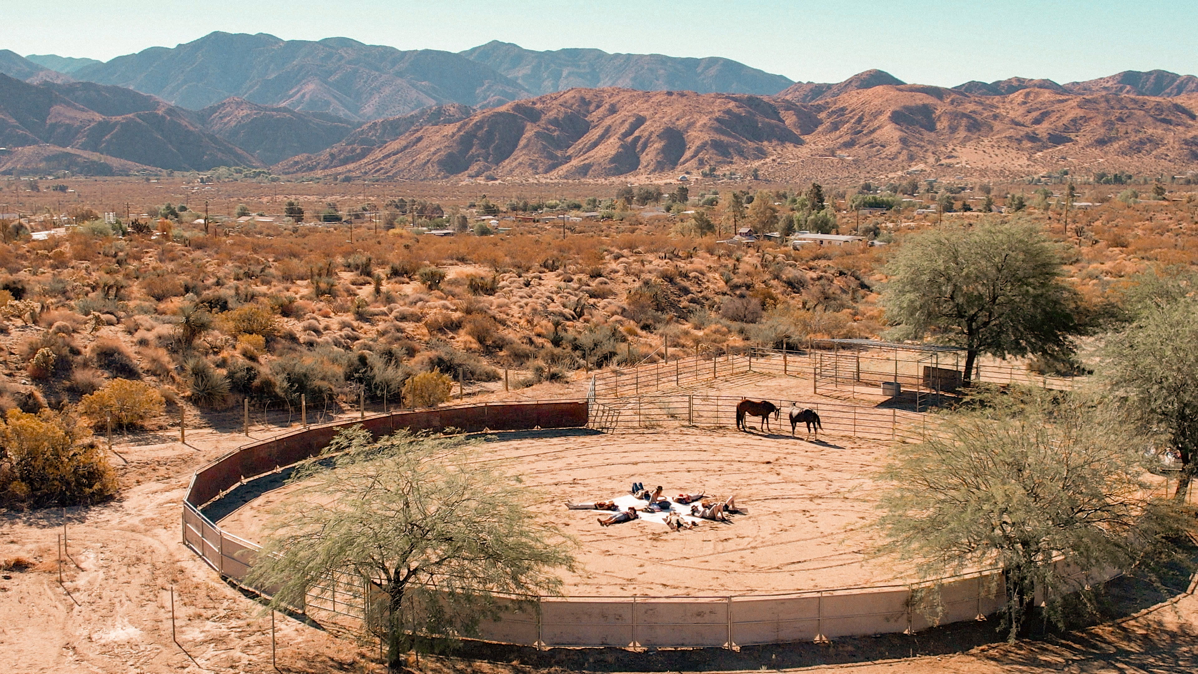 The image shows a circular horse corral surrounded by a desert landscape with mountains in the background and a few horses inside the enclosure.