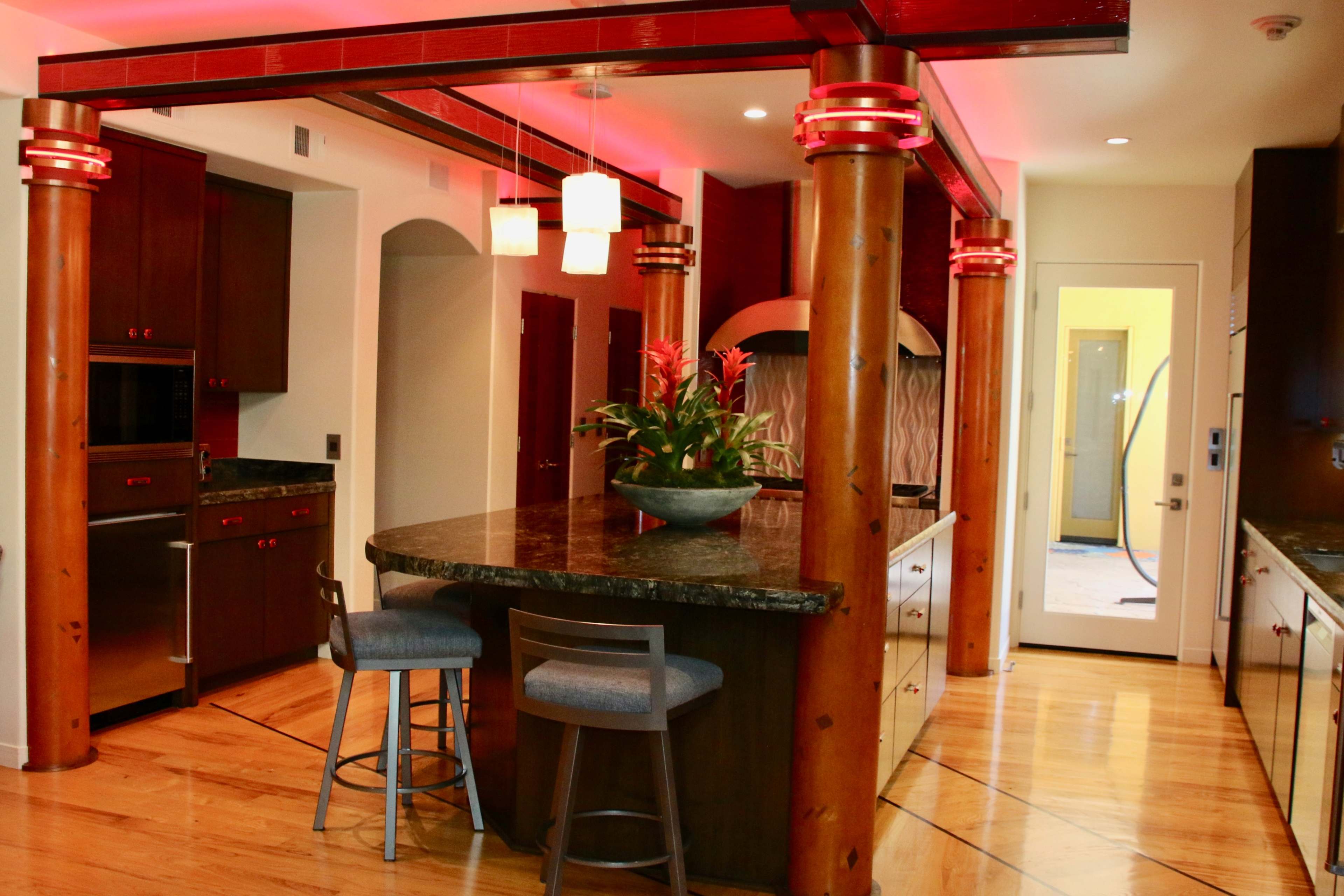 A modern kitchen with a large island, wooden pillars, and pendant lighting, featuring dark cabinetry and a granite countertop.