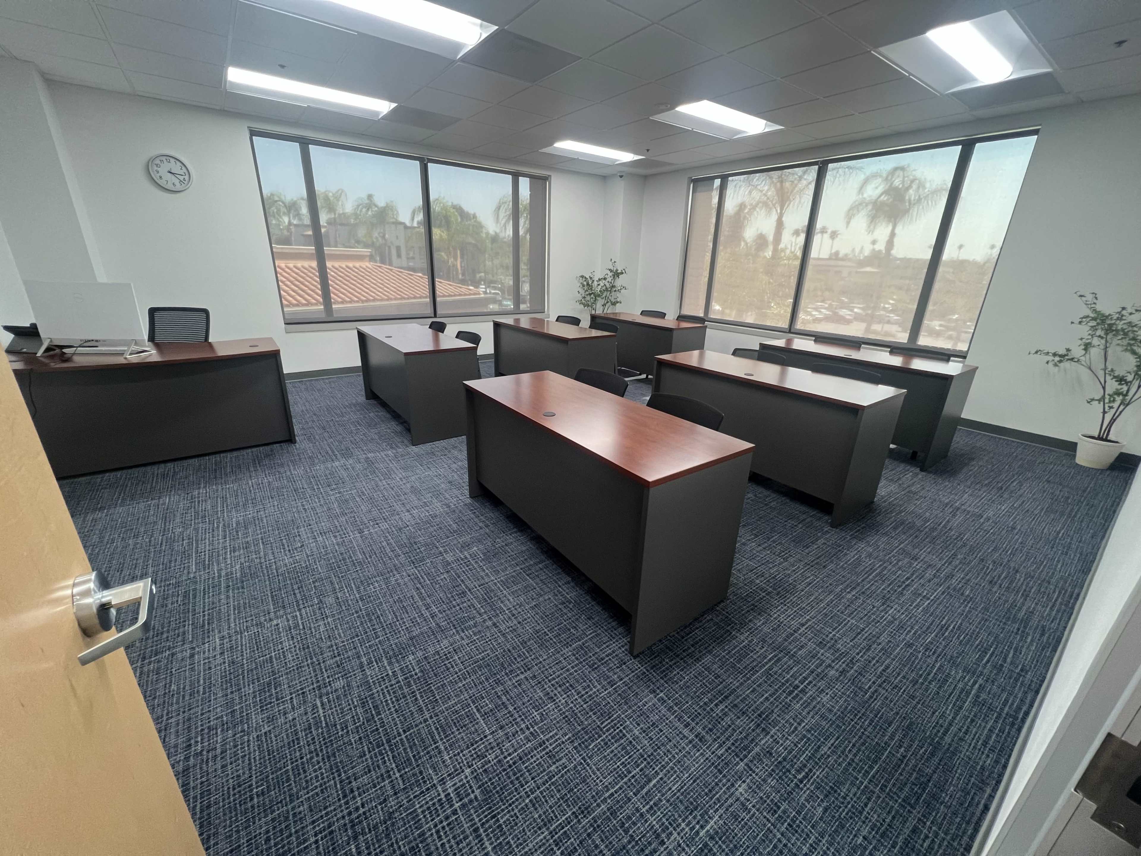 An office space features several dark wooden desks arranged in a spacious layout with large windows overlooking a palm tree view.