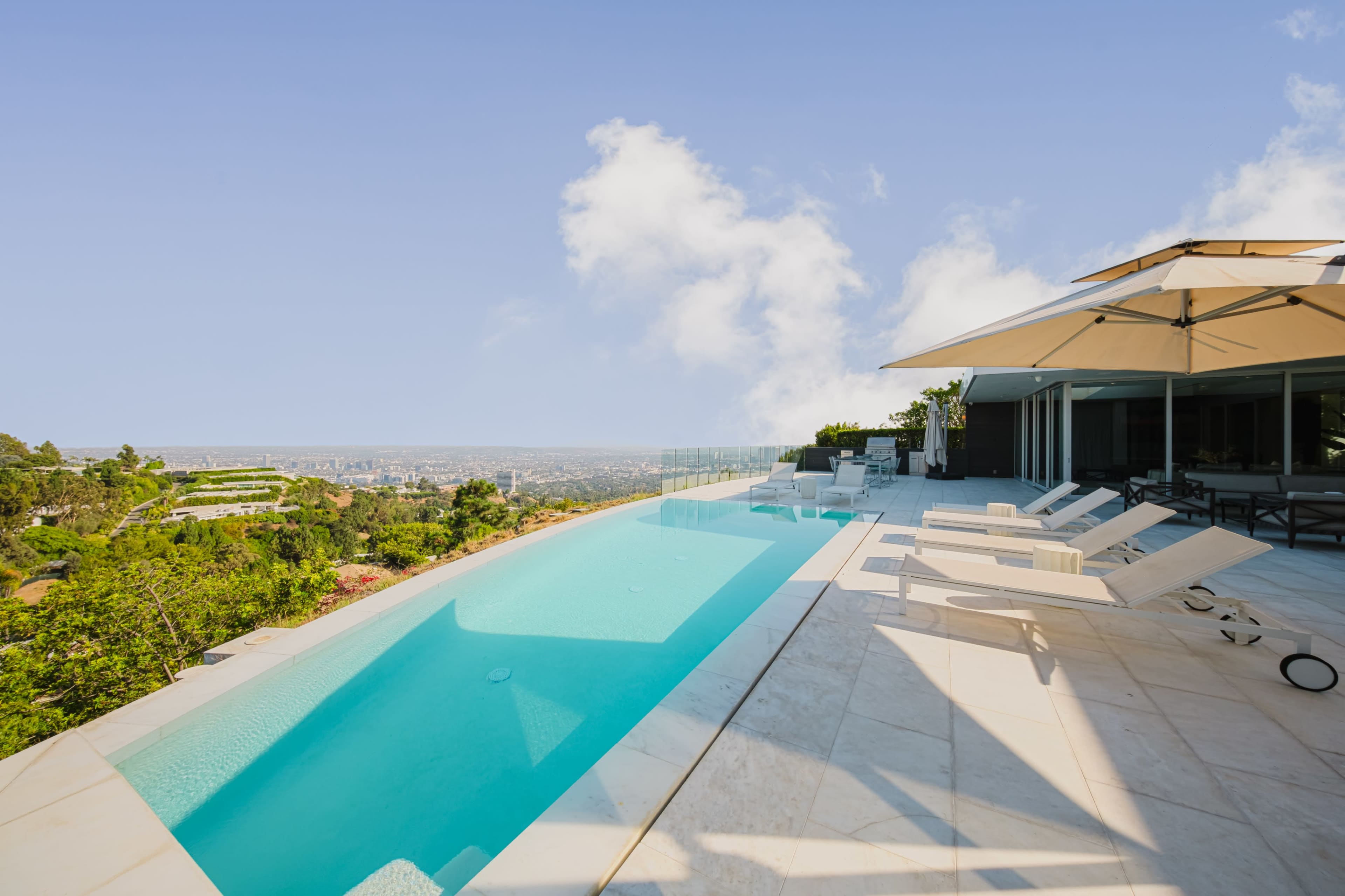 The image shows a modern villa with an infinity pool overlooking a cityscape, surrounded by greenery and blue skies.