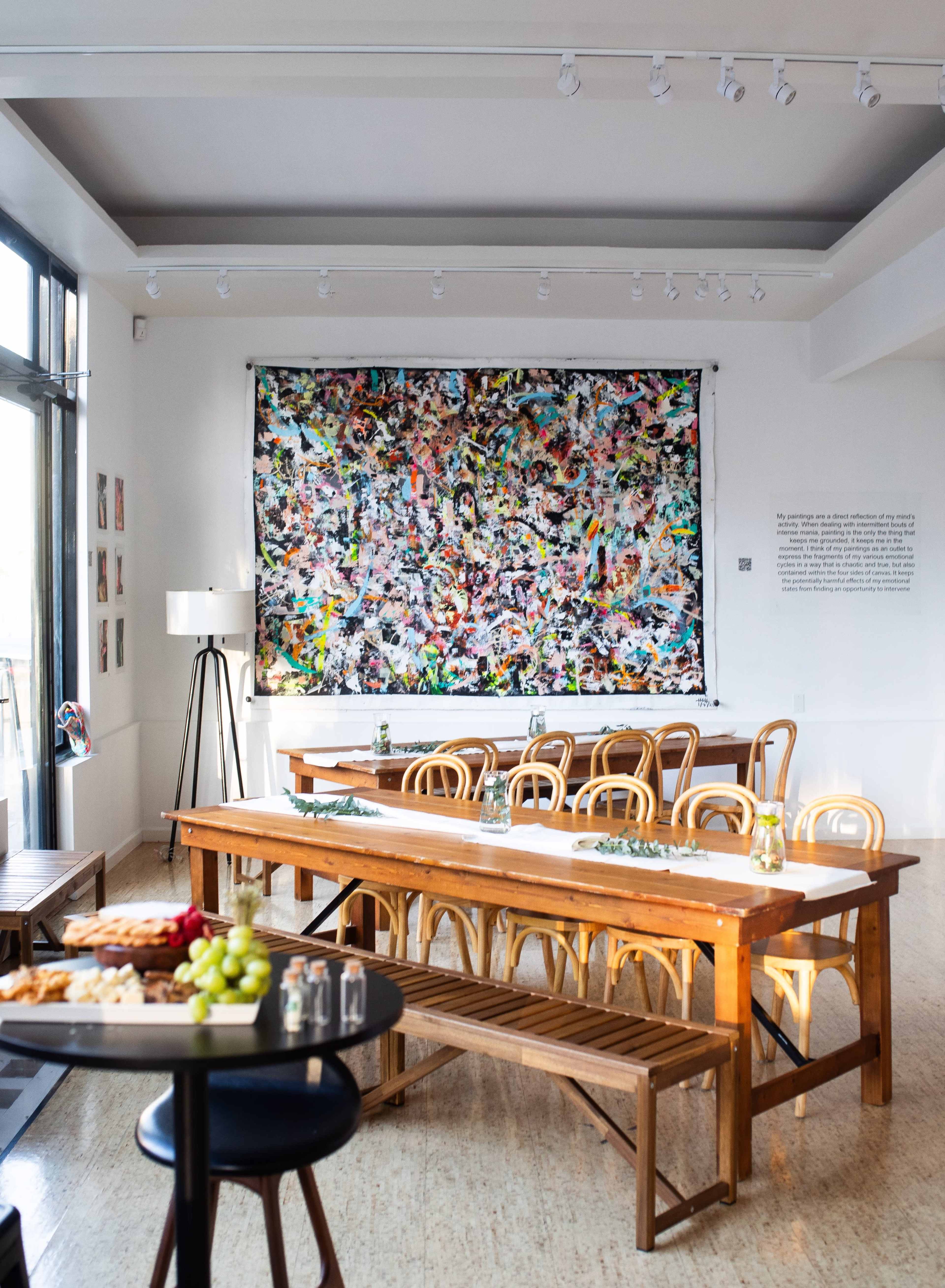 A spacious dining area features long wooden tables set with small vases, a large colorful abstract painting on the wall, and a side bar holding food and drinks.