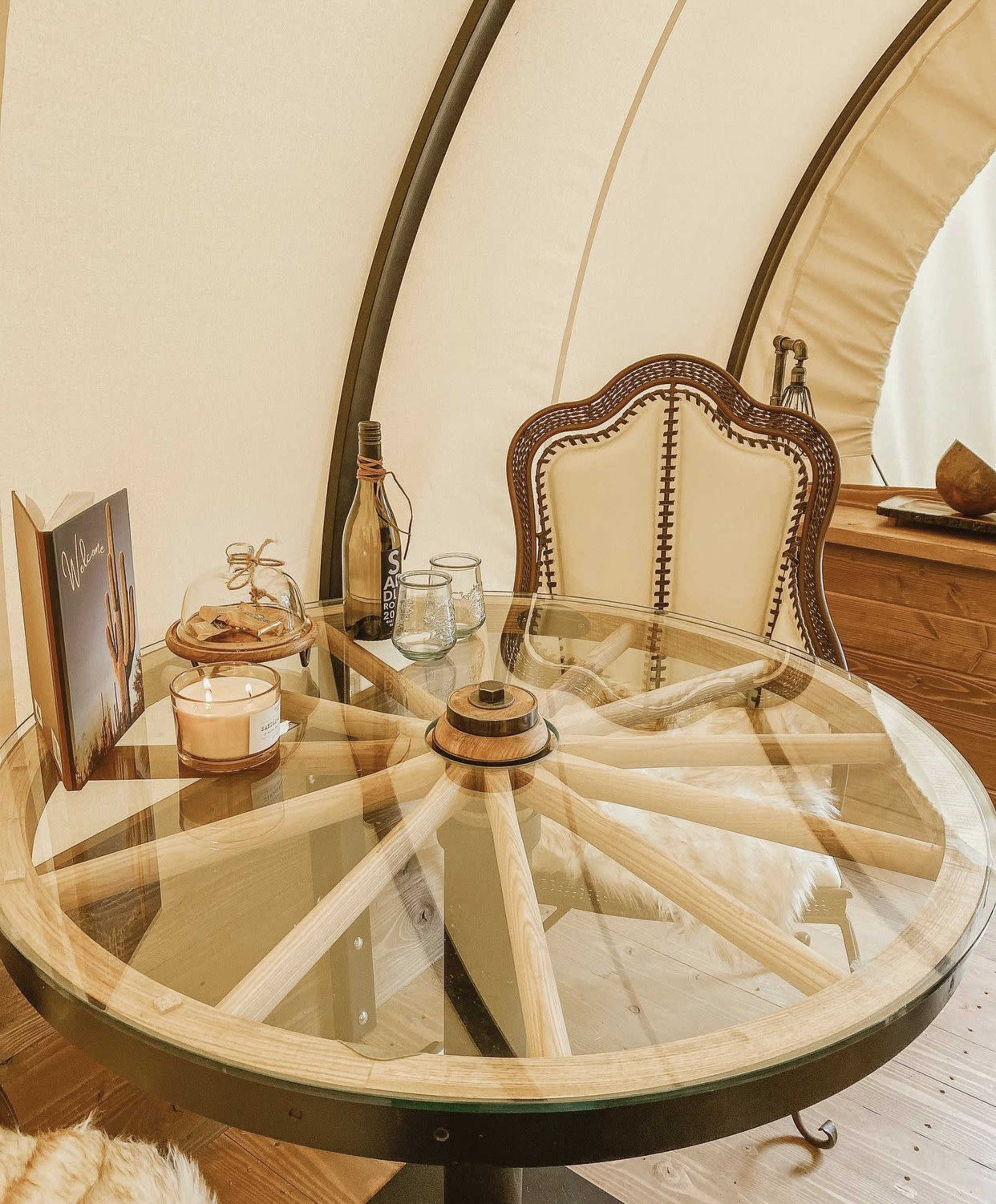 A glass-top table with a wooden wheel design, accompanied by a decorative chair, a candle, a bottle, and a book on a nearby surface inside a cozy setting.