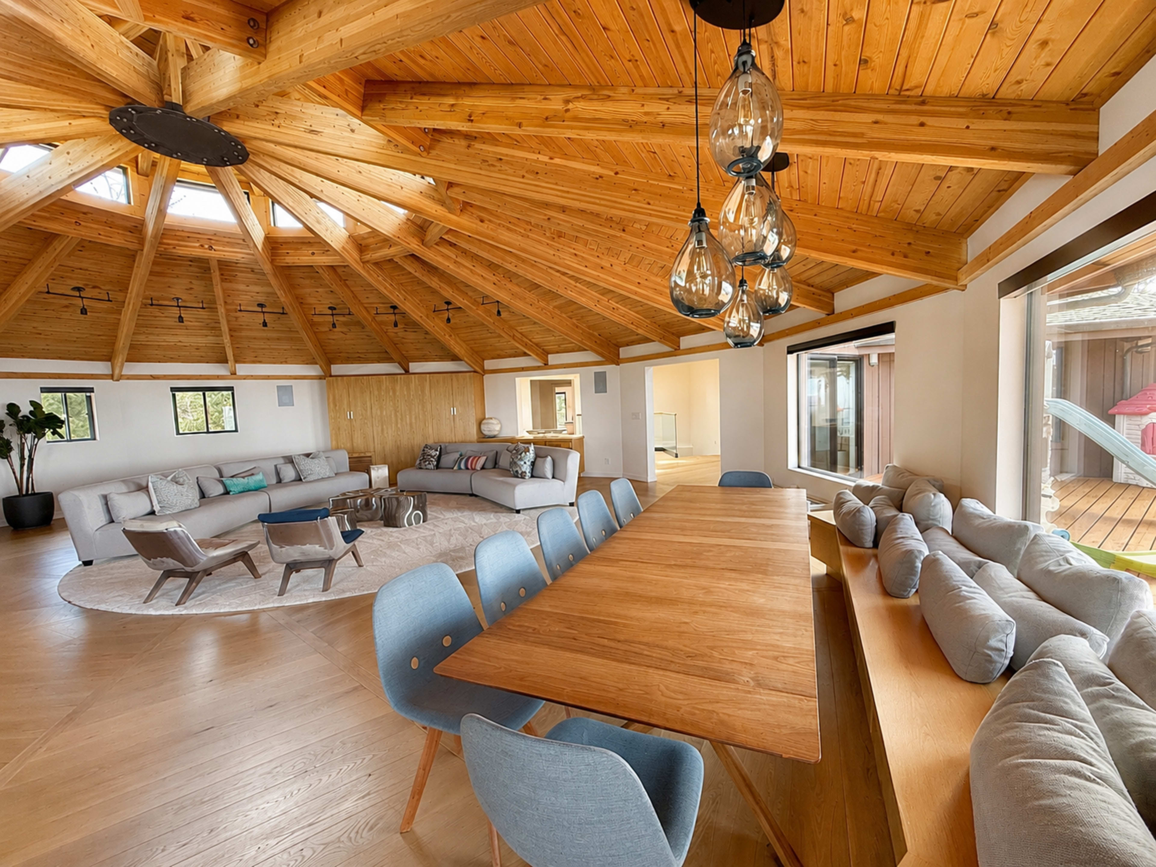 The image shows a spacious, circular living area with a high, wooden, vaulted ceiling, featuring a dining table and various seating arrangements.