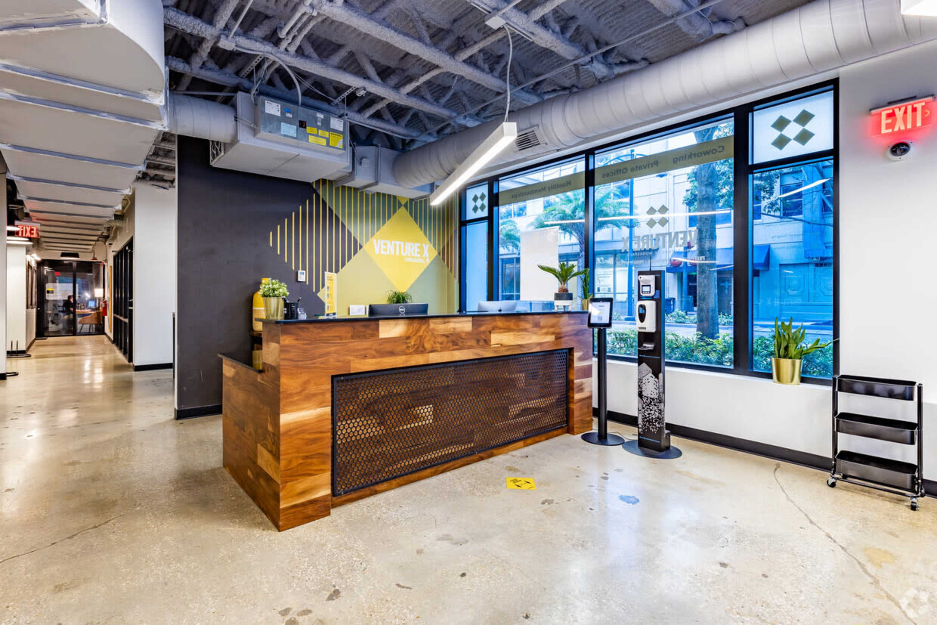 A modern reception area with a wooden front desk, large windows, and plants near the windows.