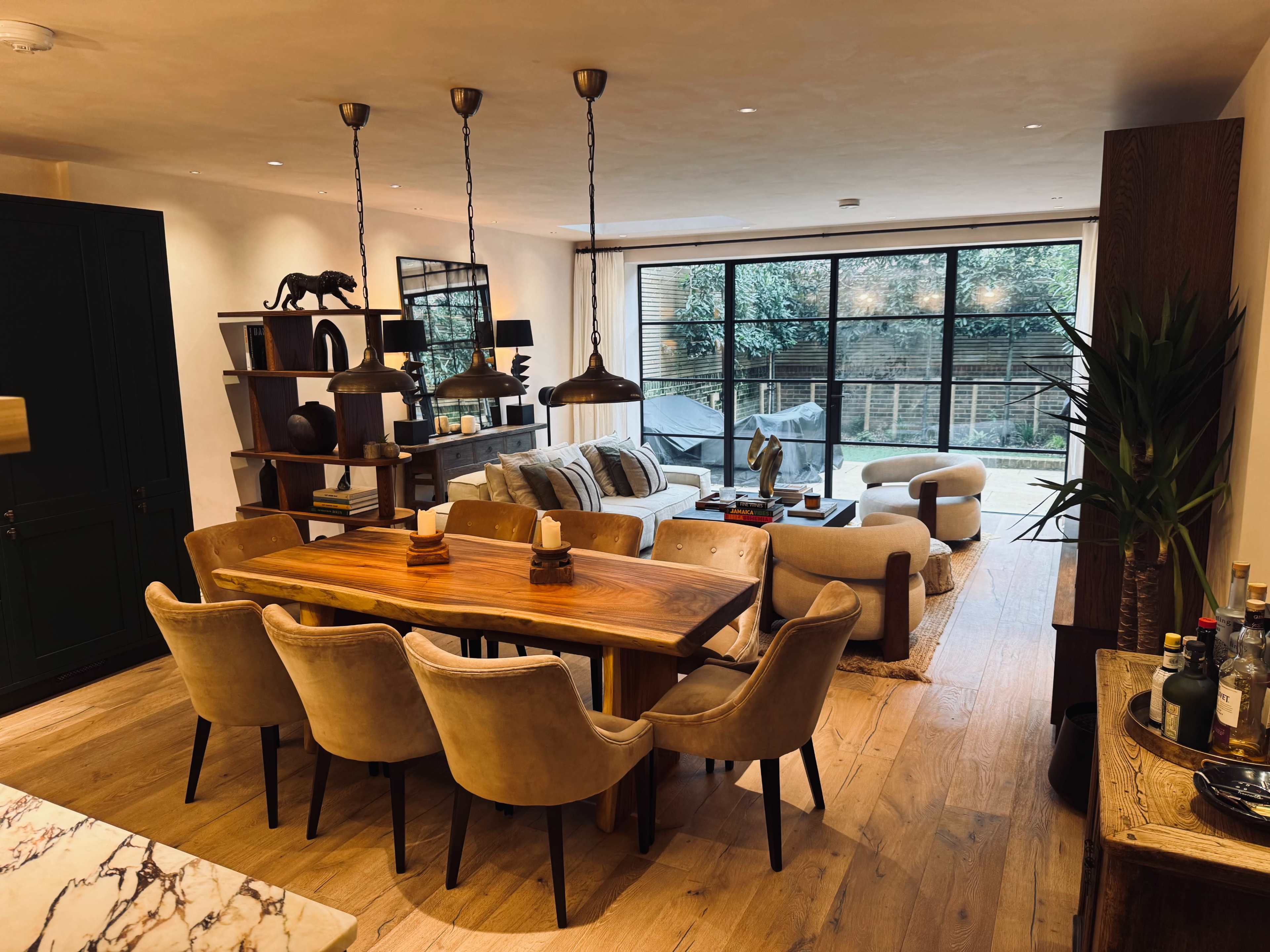 A dining area featuring a wooden table surrounded by upholstered chairs, adjacent to a living space with a sofa and large windows overlooking a garden.