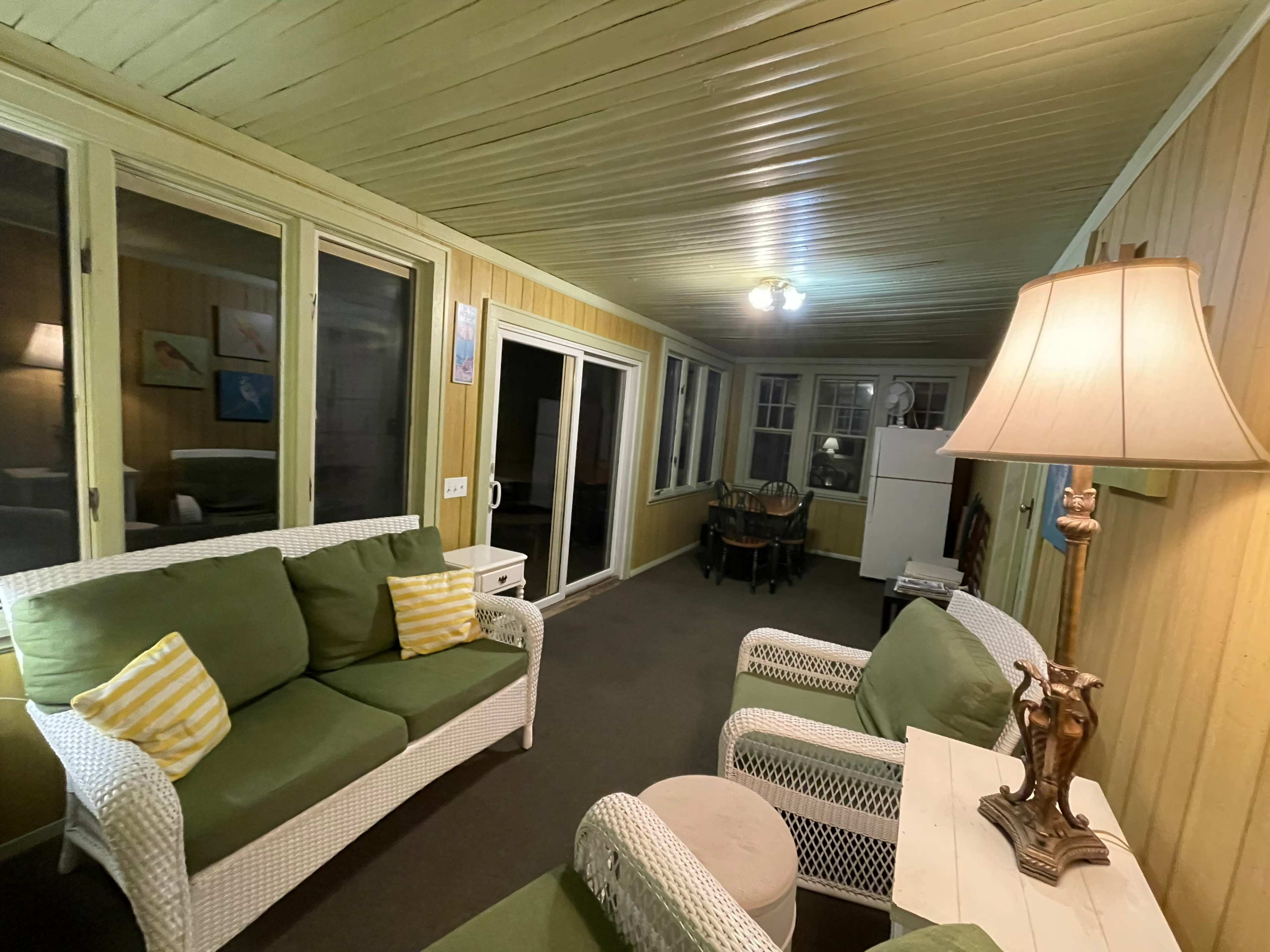 A sunroom with green sofas, a white lamp, and a dining area visible in the background.