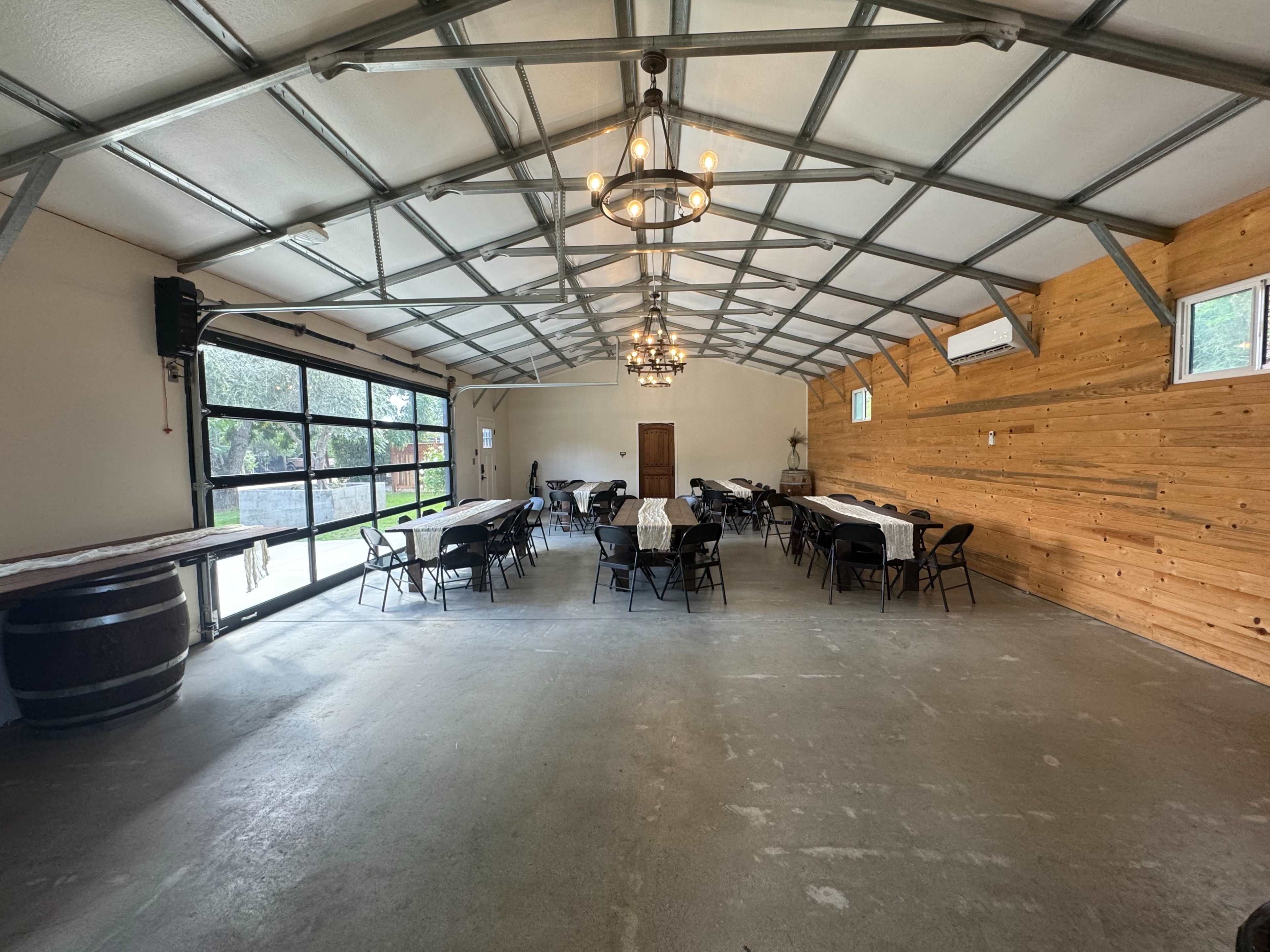 The image shows a spacious, well-lit indoor venue with rows of tables and chairs, a wooden accent wall, and large glass doors leading outside.