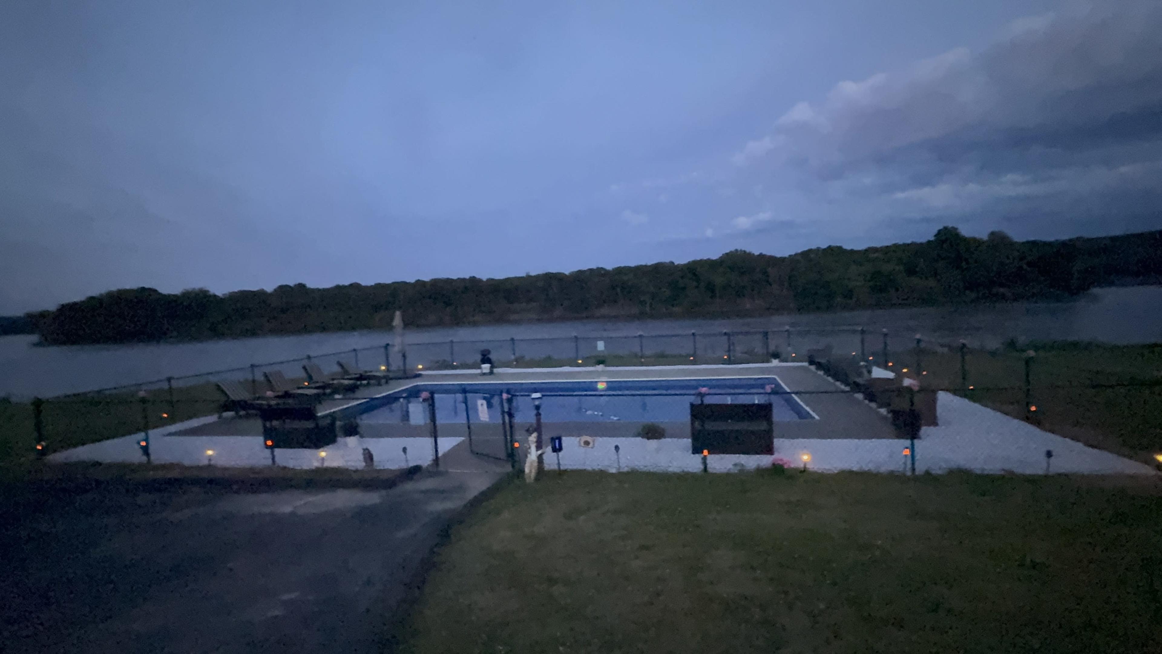 The image shows a swimming pool area beside a river, surrounded by a grassy lawn and a fence, under an evening sky.