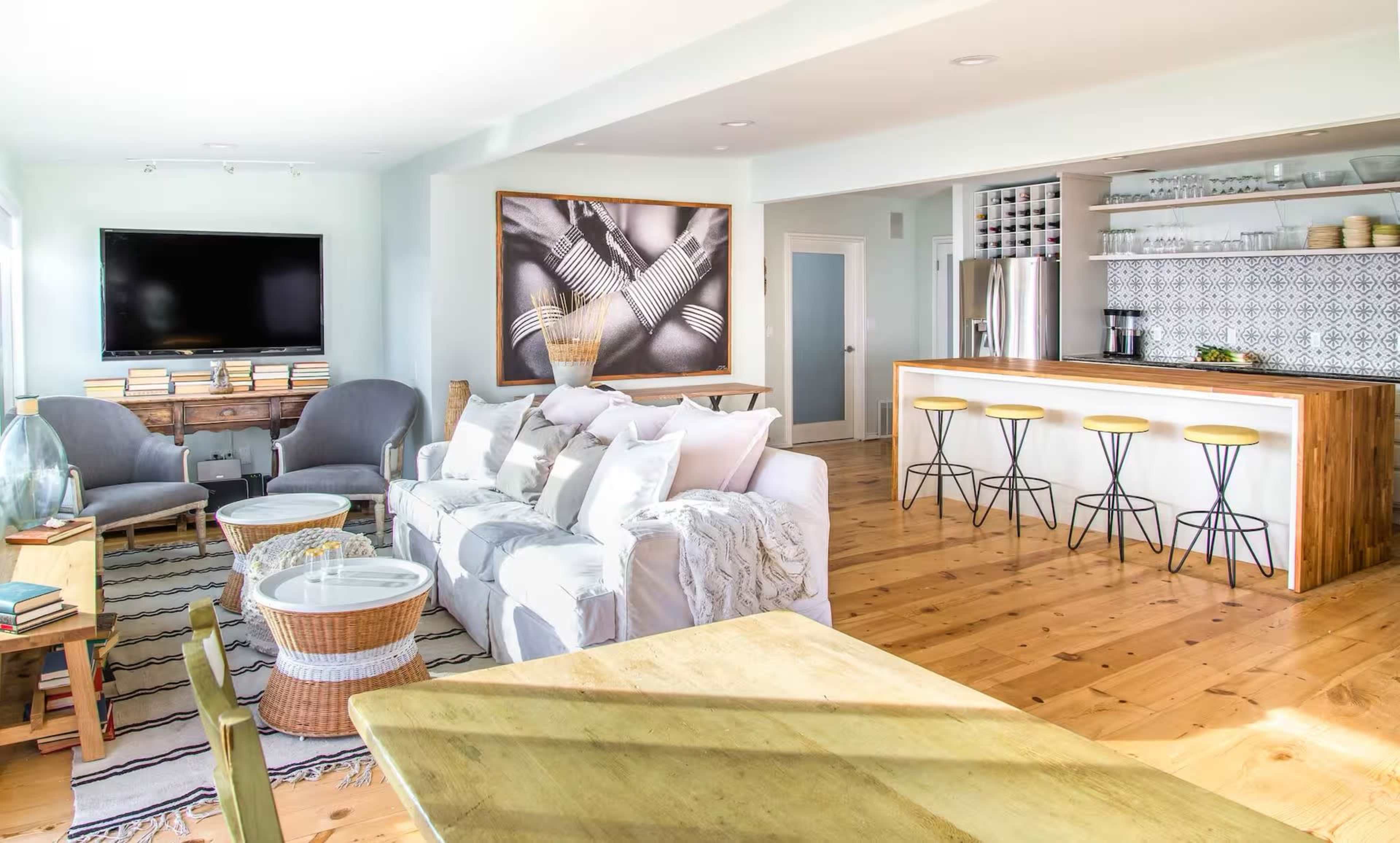 A modern open-concept living area features a comfortable seating arrangement, a kitchen with bar stools, and a large artwork on the wall.