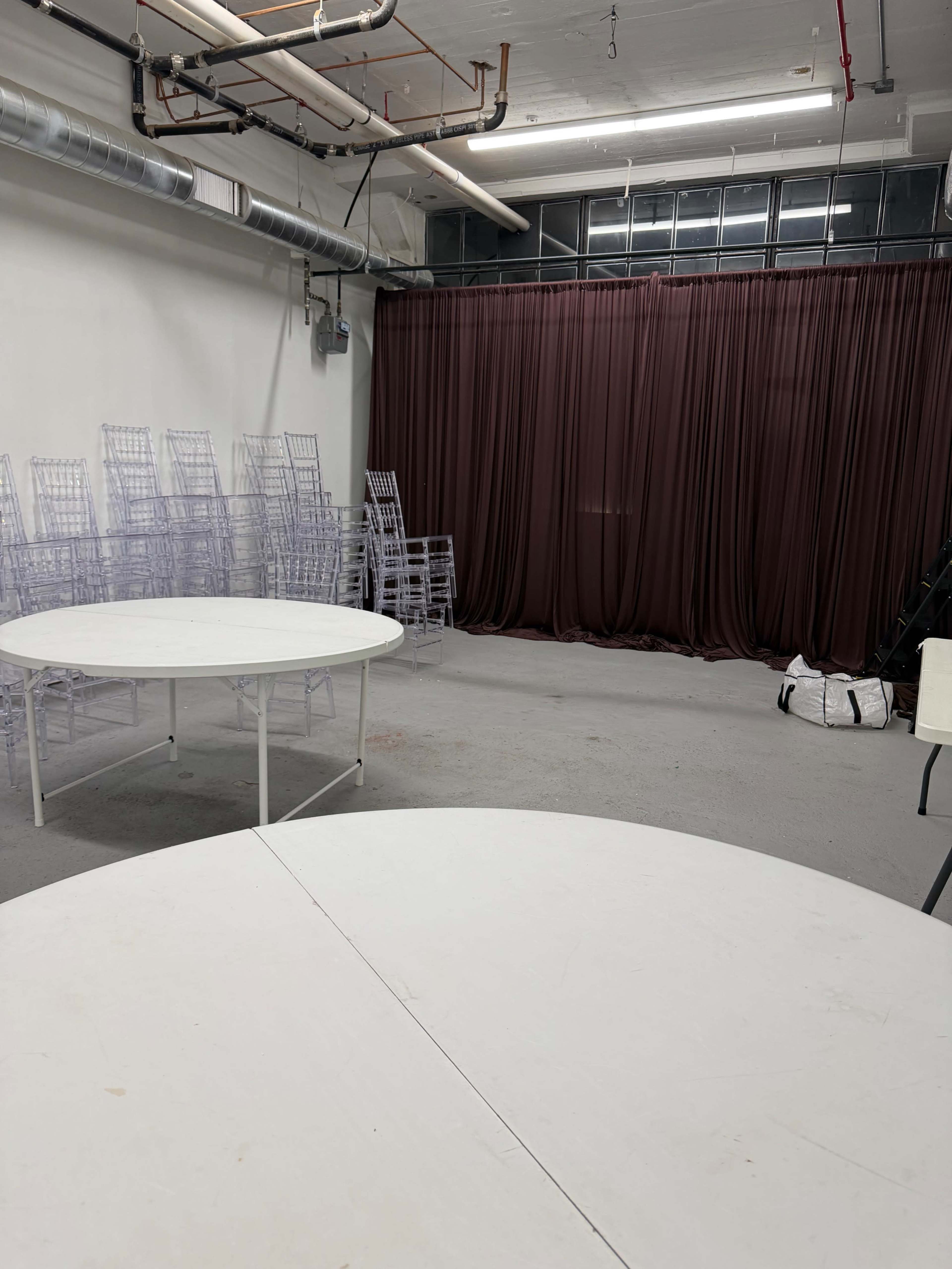 The image shows an empty room with two white tables in the foreground, a stack of clear chairs behind them, and a curtain in the background.