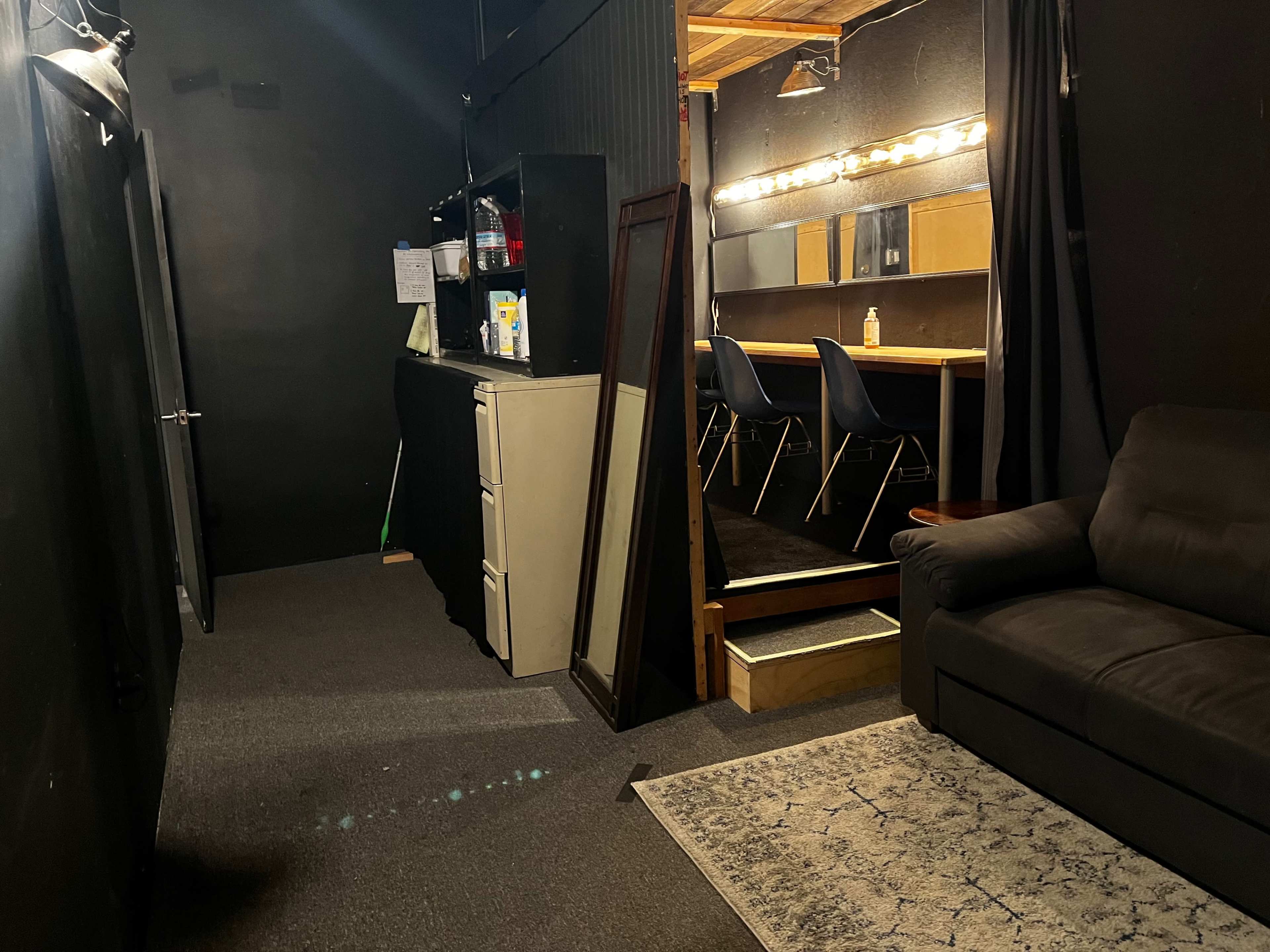 The scene shows a dark hallway leading to a well-lit area with a mirror and a row of chairs, alongside a couch and a cabinet.