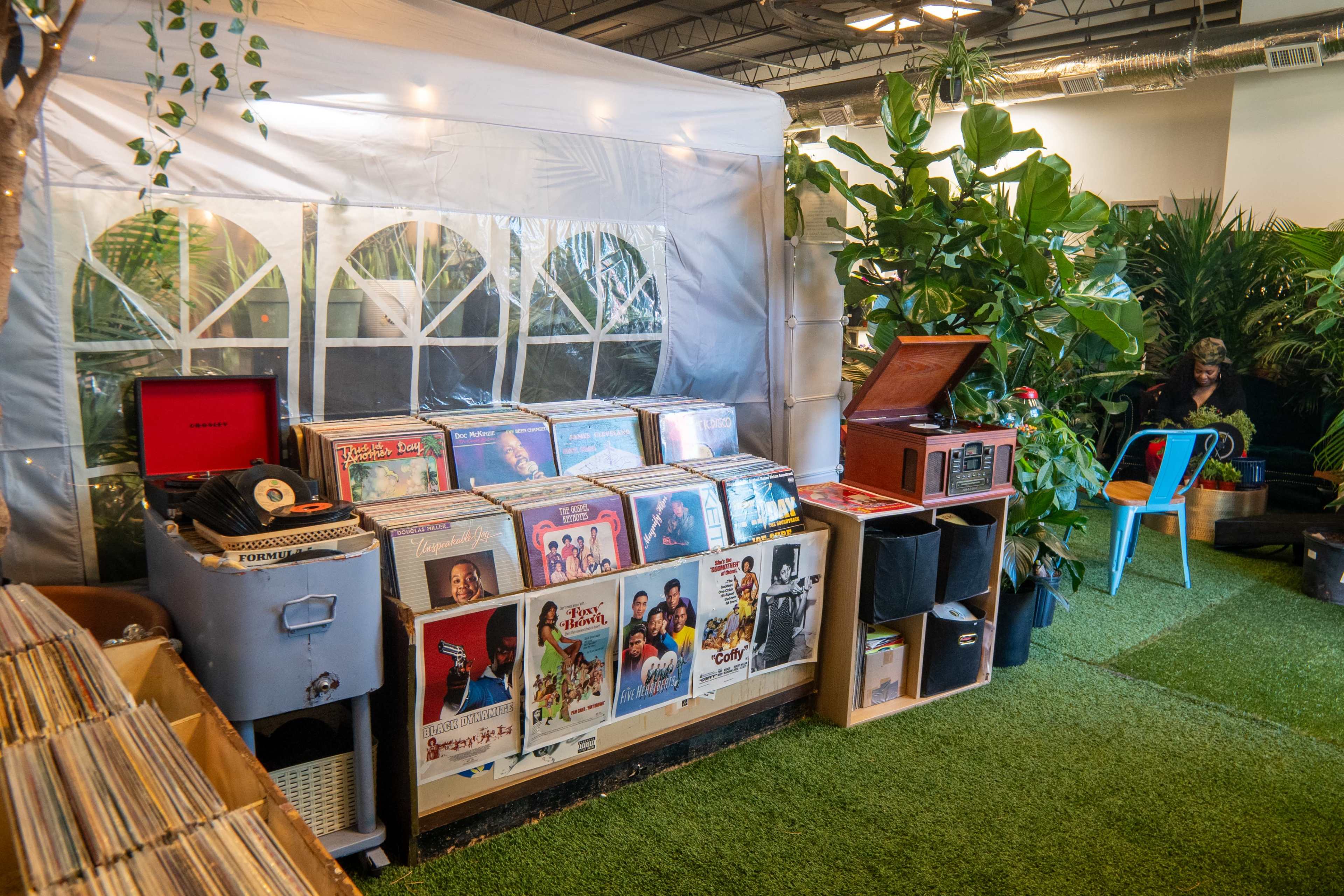 The image shows a collection of vinyl records displayed on shelves alongside a vintage record player, set in a green, plant-filled environment with a tent-like structure in the background.