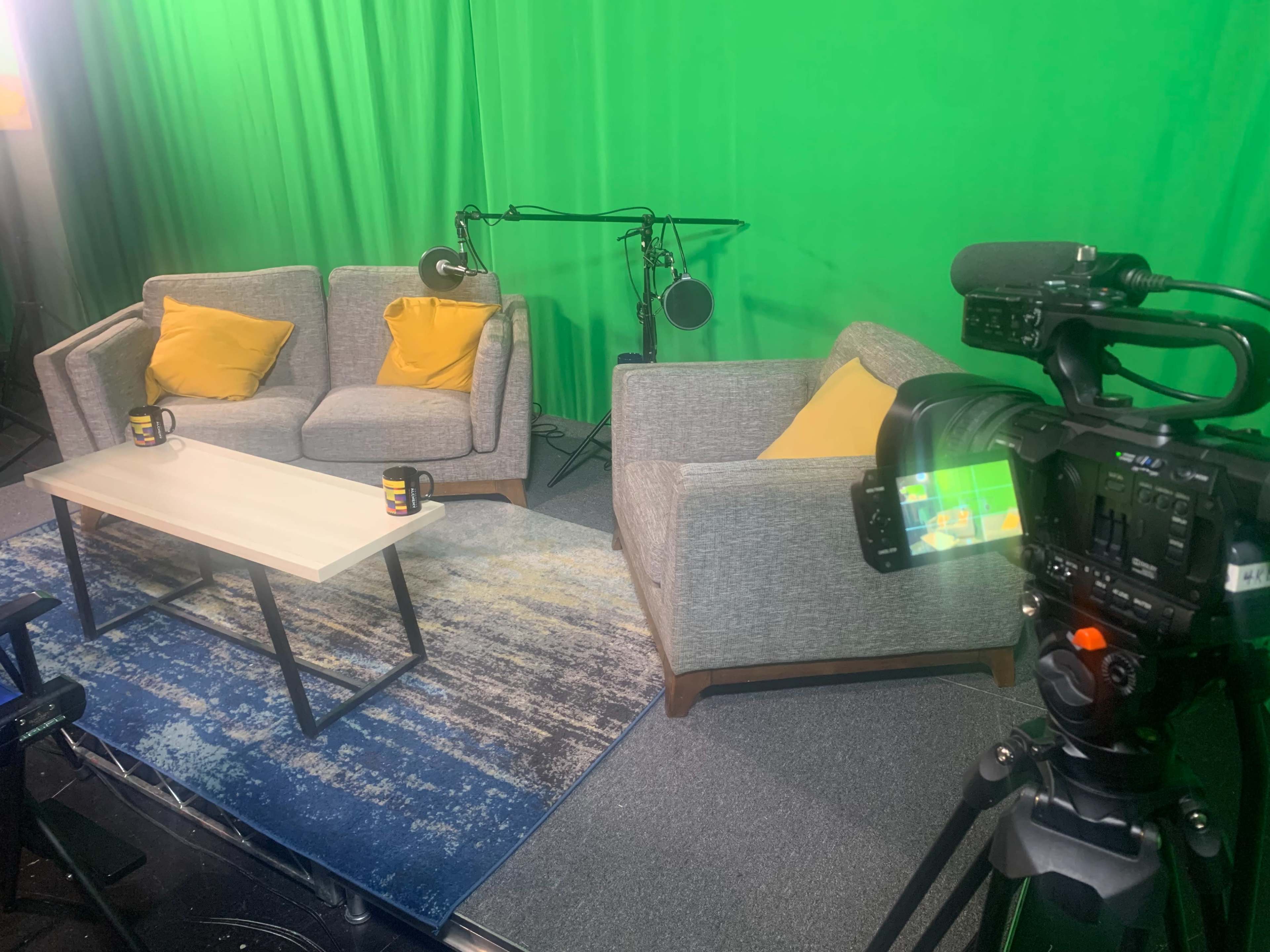 A camera points toward a set with two gray couches, yellow pillows, and a white coffee table in front of a green screen.