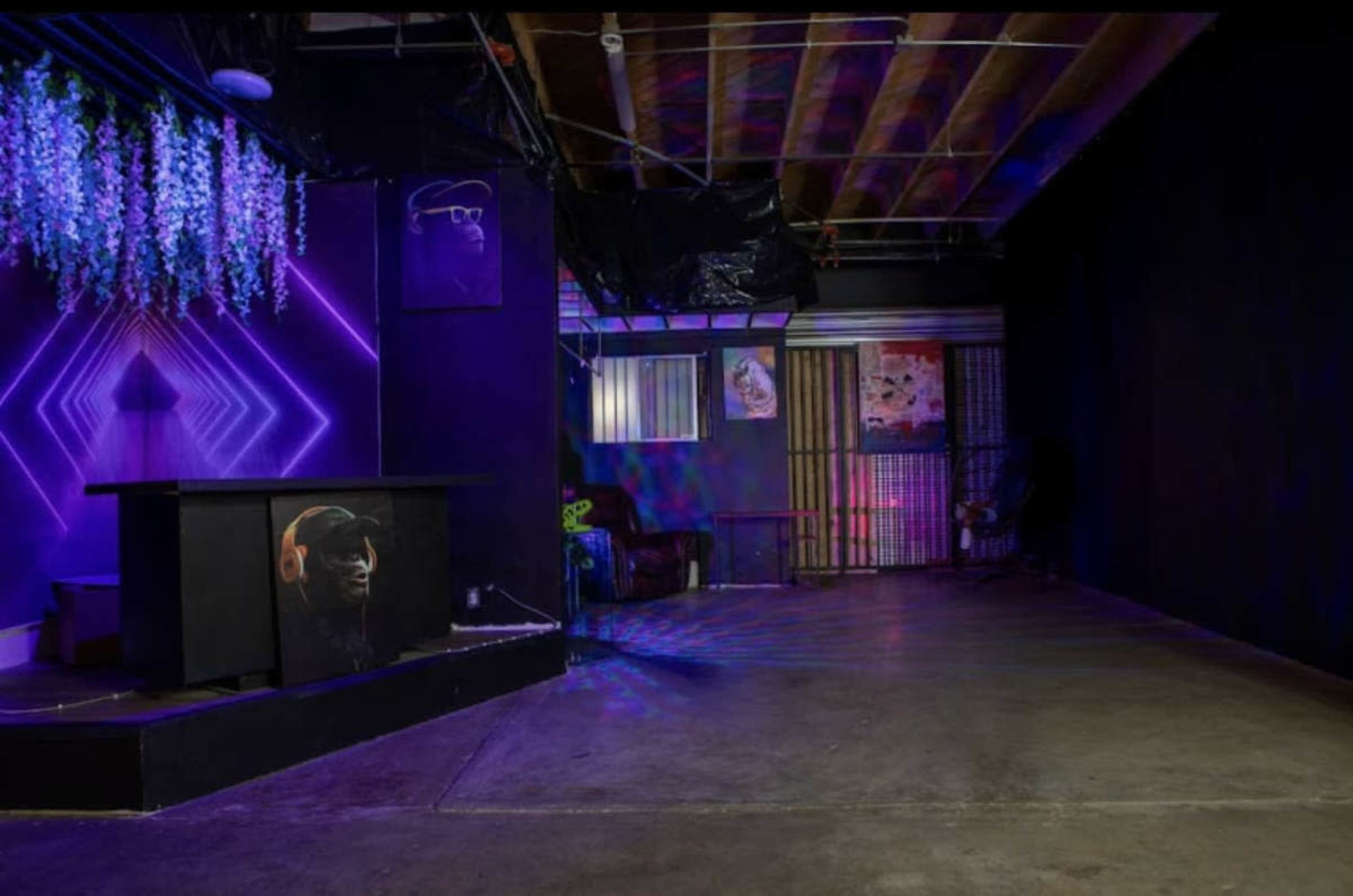 The image shows a dimly lit room with a decorative bar featuring purple lighting, surrounded by sparse furniture and walls painted in dark colors.