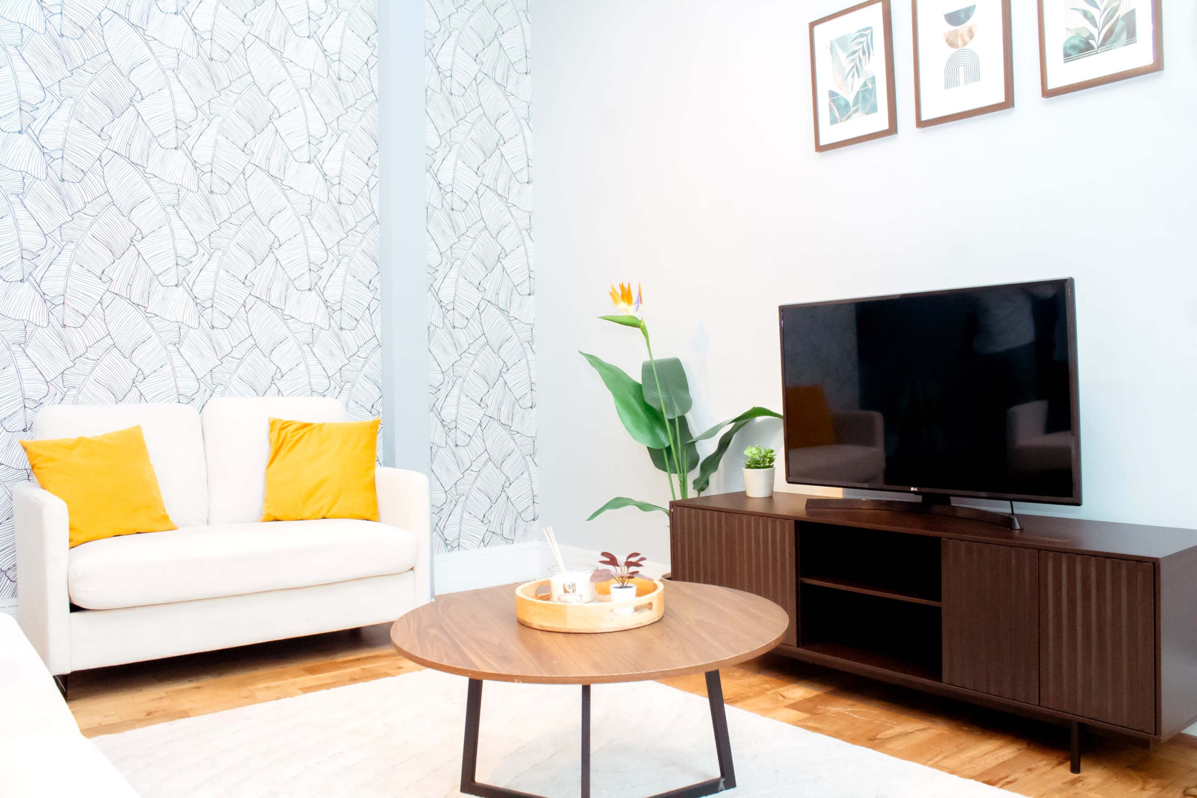 The image shows a modern living room with a white sofa, a round wooden coffee table, a television on a dark wood stand, and decorative wall art.