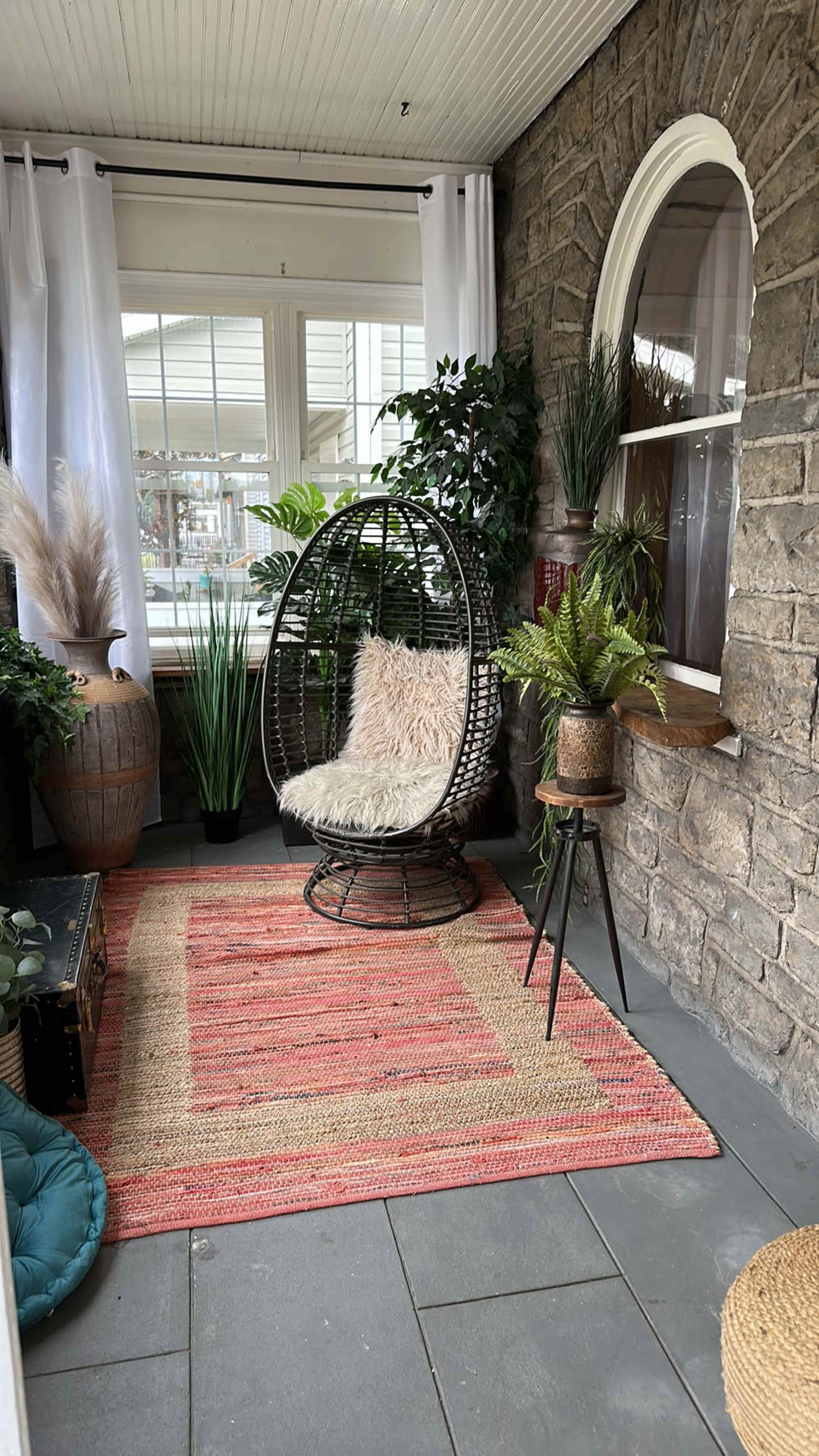 A cozy sunroom features a hanging chair, plants, and a woven rug on a stone floor.