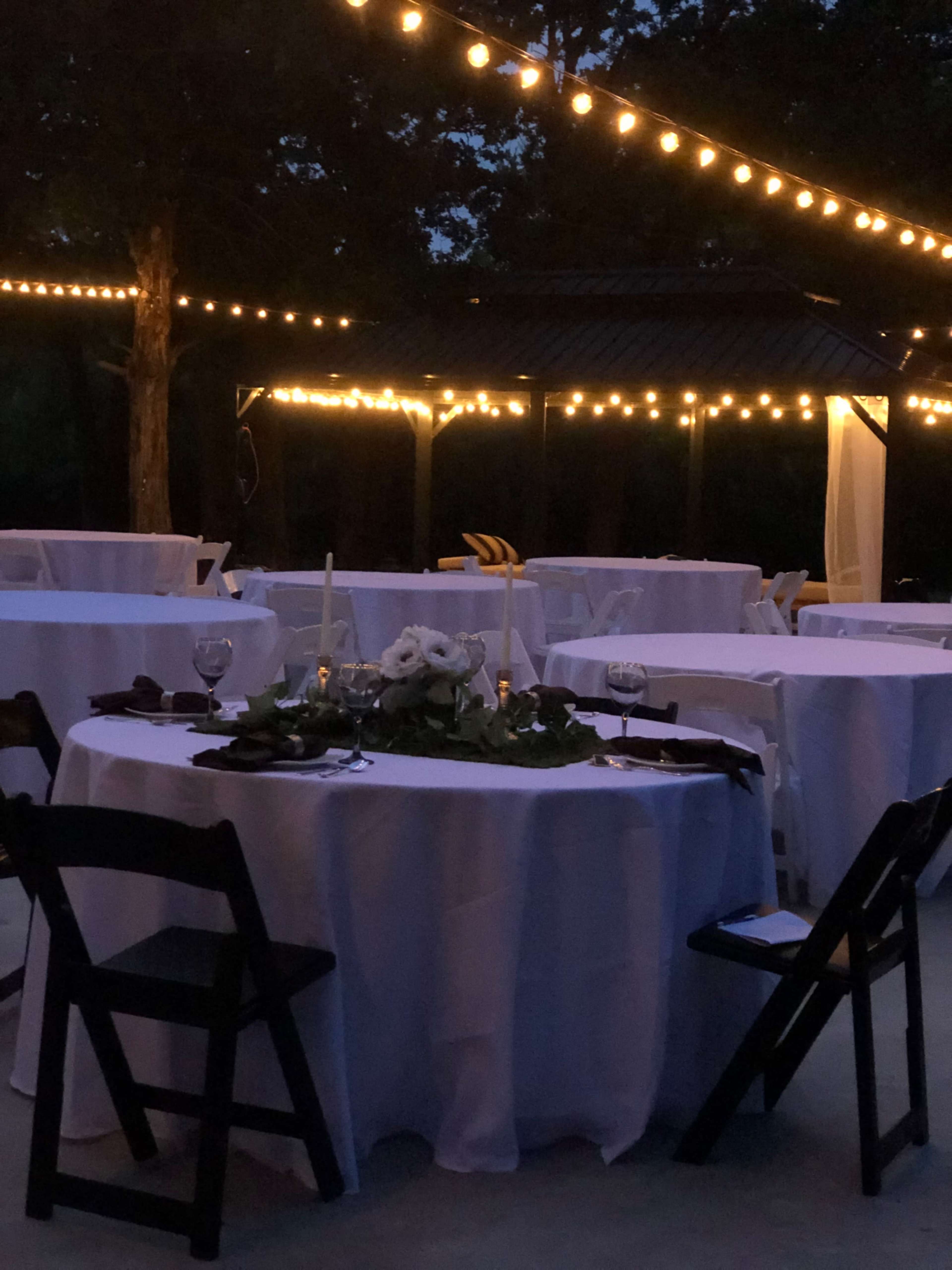 The scene features several round tables set with white tablecloths and surrounded by black chairs, illuminated by string lights in a dimly lit outdoor setting.