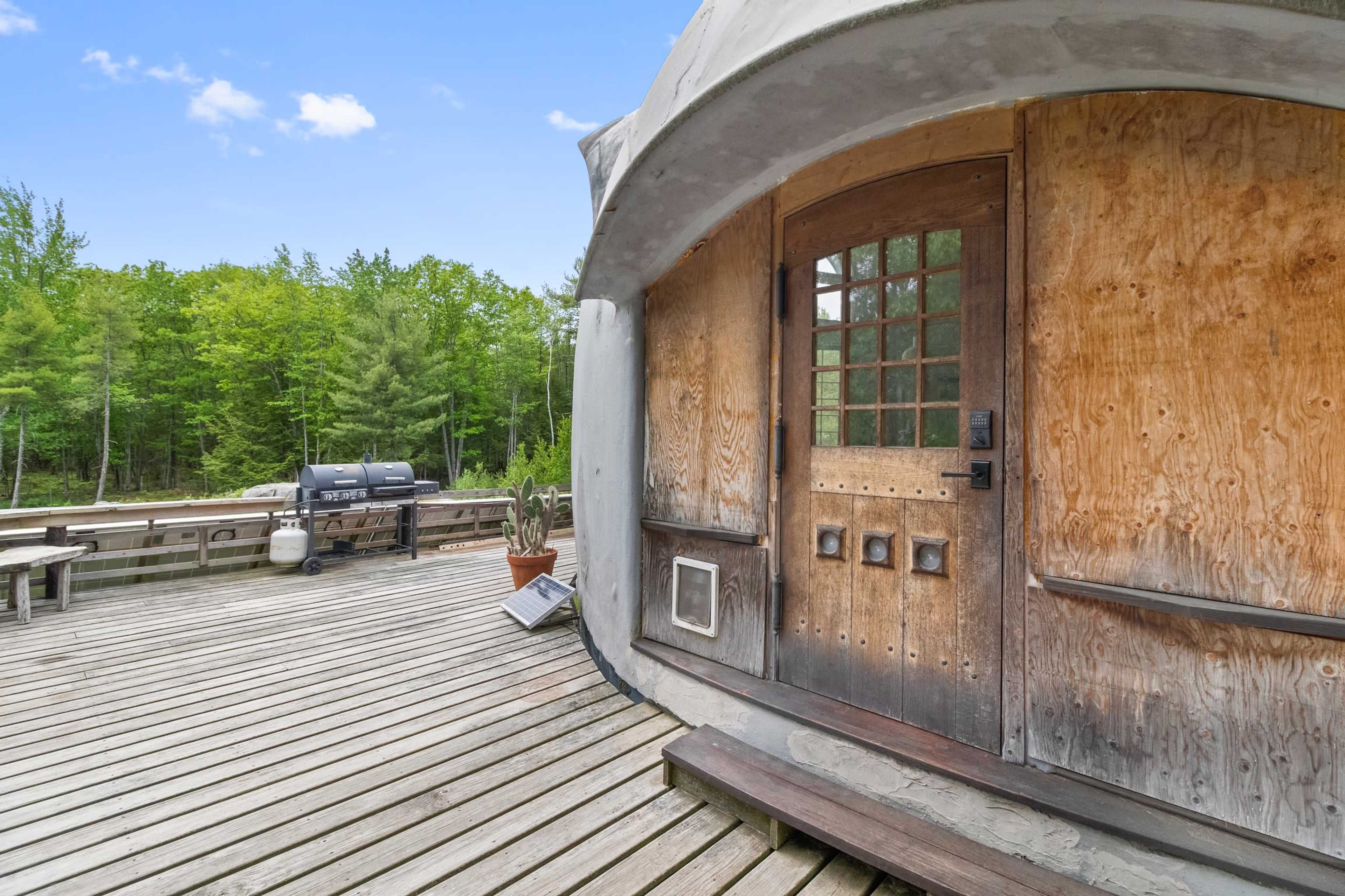 A wooden deck leads to a rounded entrance of a cabin, surrounded by trees and featuring a grill in the background.