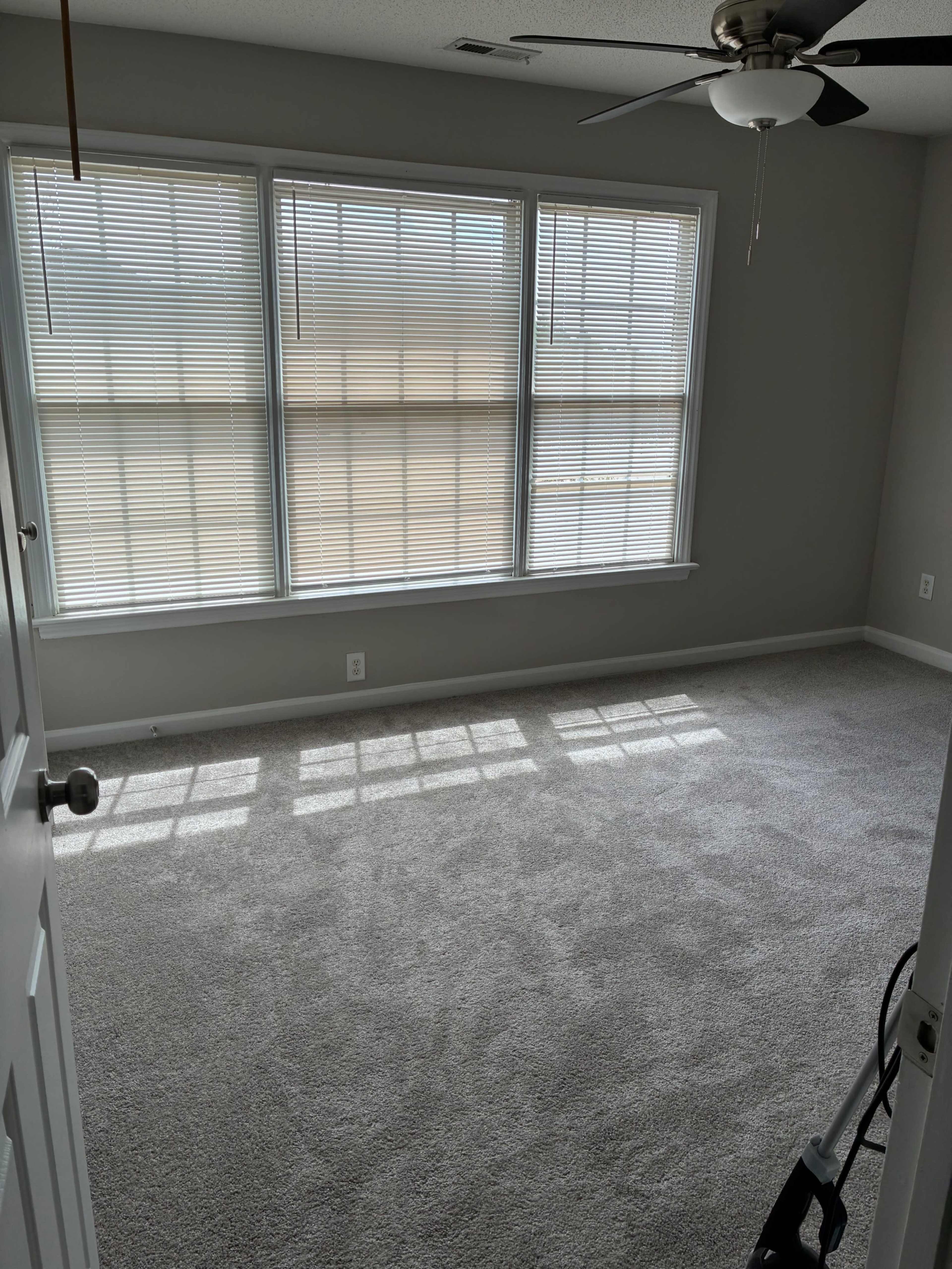 The image shows a vacant room with light gray walls, a carpeted floor, and three large windows allowing natural light to enter.