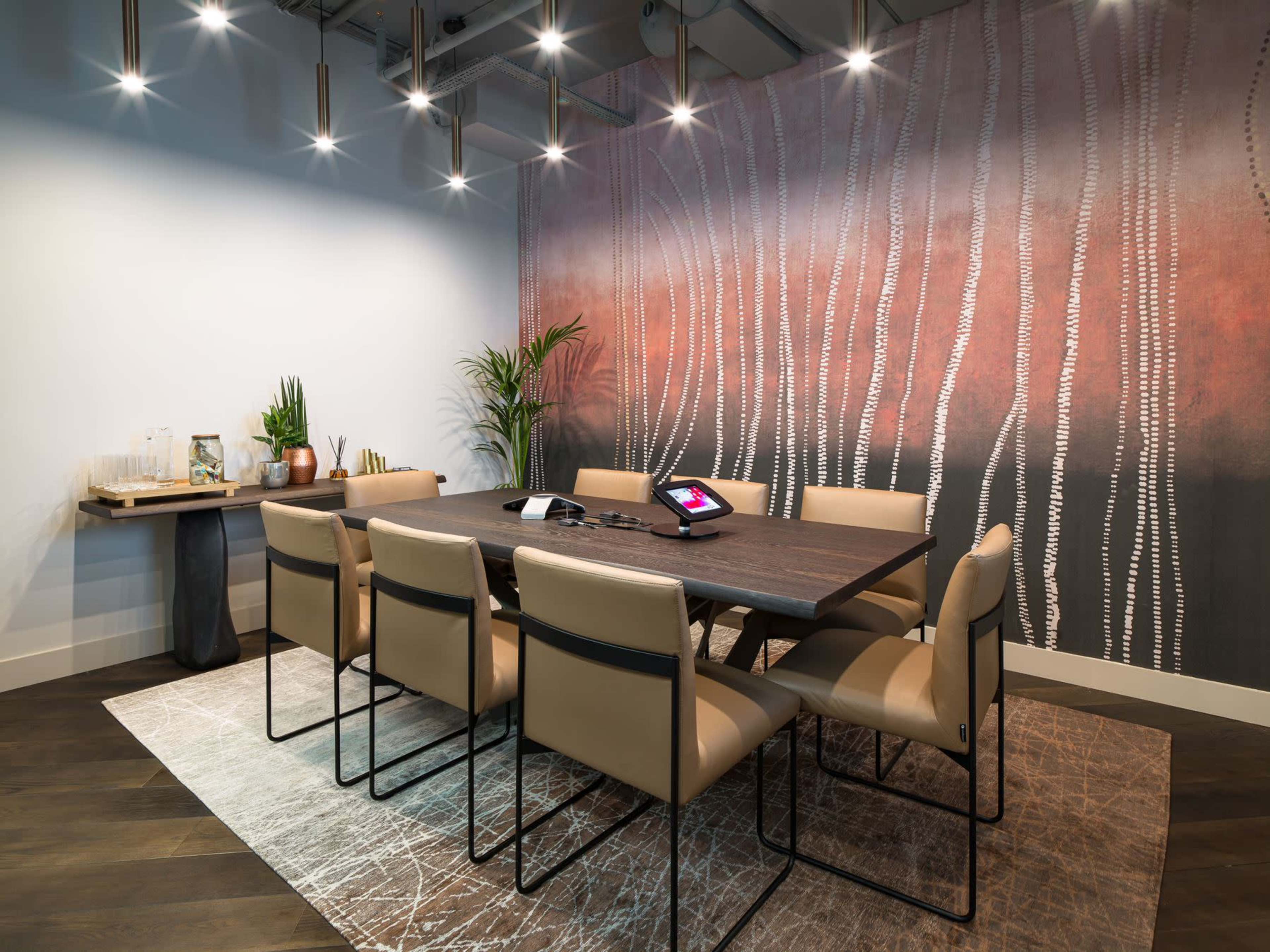 A modern conference room features a large wooden table surrounded by beige chairs, with a decorative wall mural and plants adding visual interest.