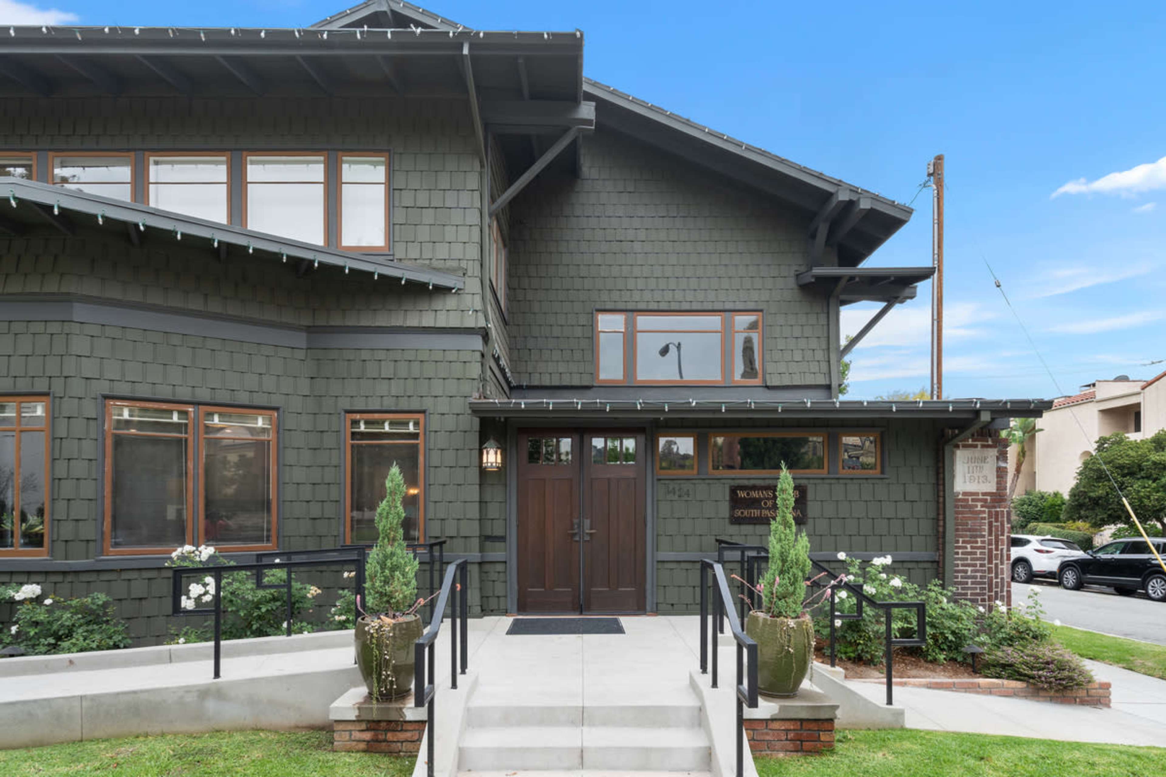 The image shows the entrance of a green, multi-story building with a wooden door, large windows, and potted plants on either side of the steps.