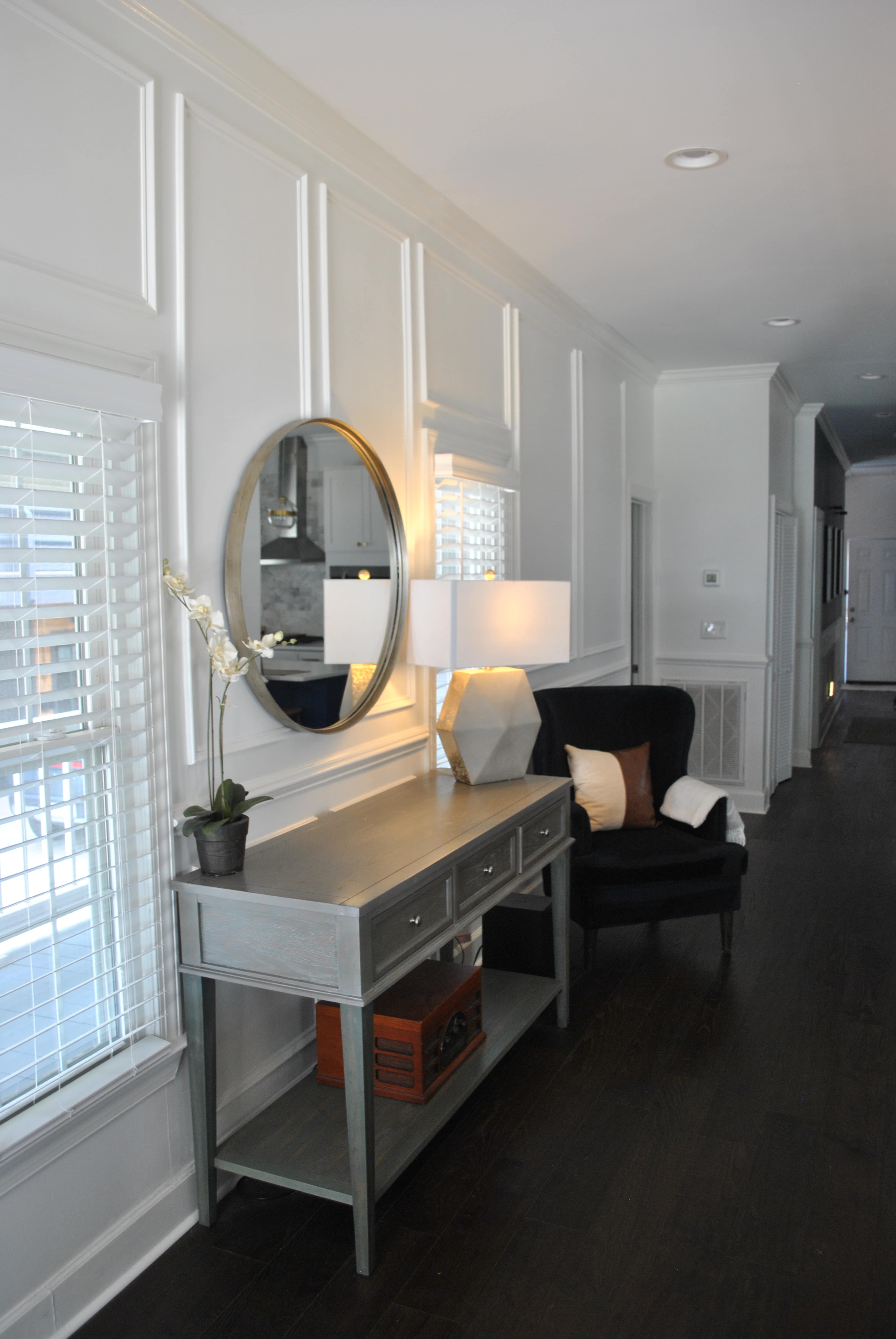 A hallway features a table with drawers, a lamp, and a round mirror, alongside a black chair and windows with white shutters.