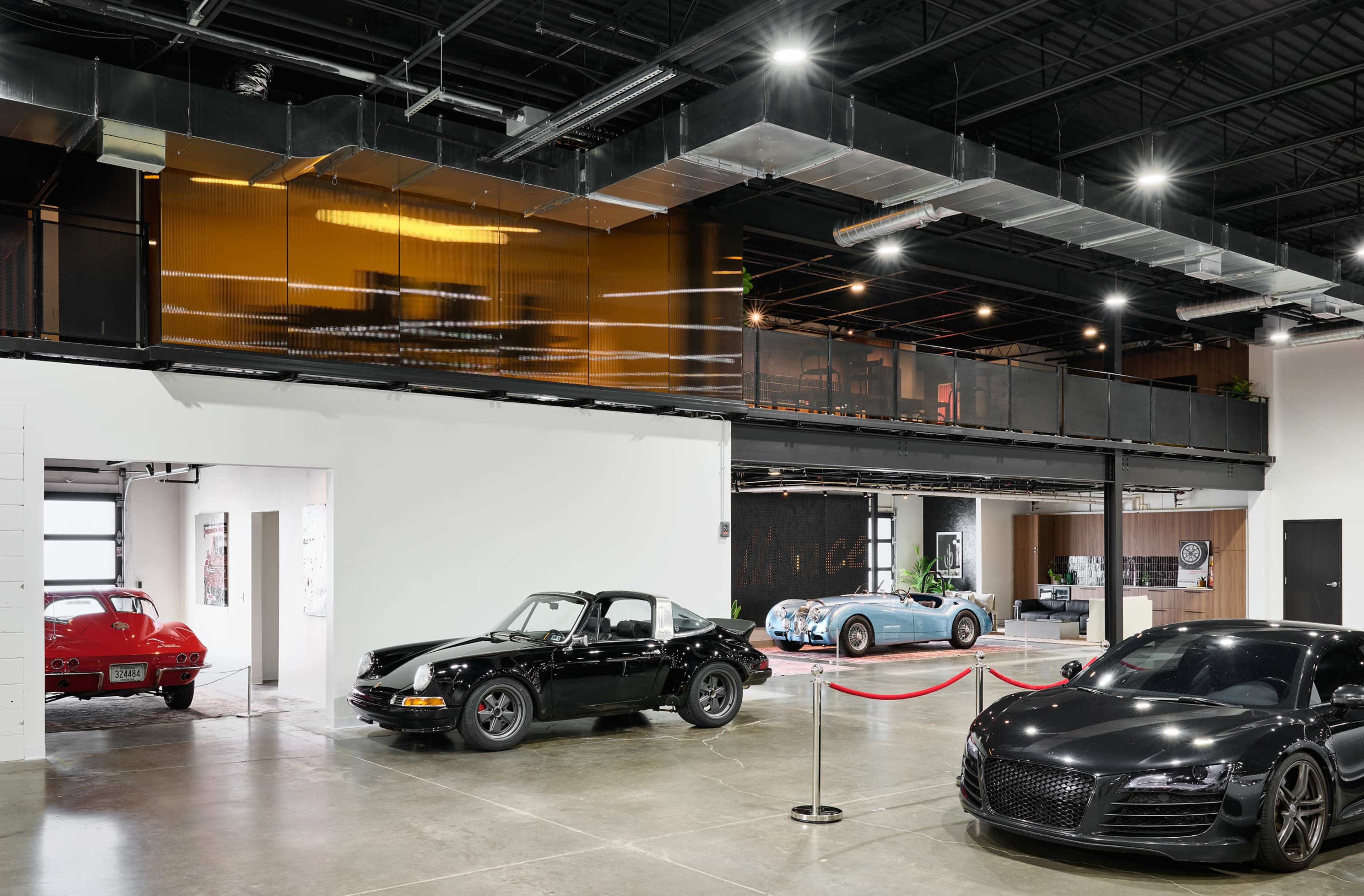 The image shows a modern garage or showroom featuring several luxury cars, including a black convertible, a red sports car, and a blue classic car, with a large open space and a glass-enclosed upper level.