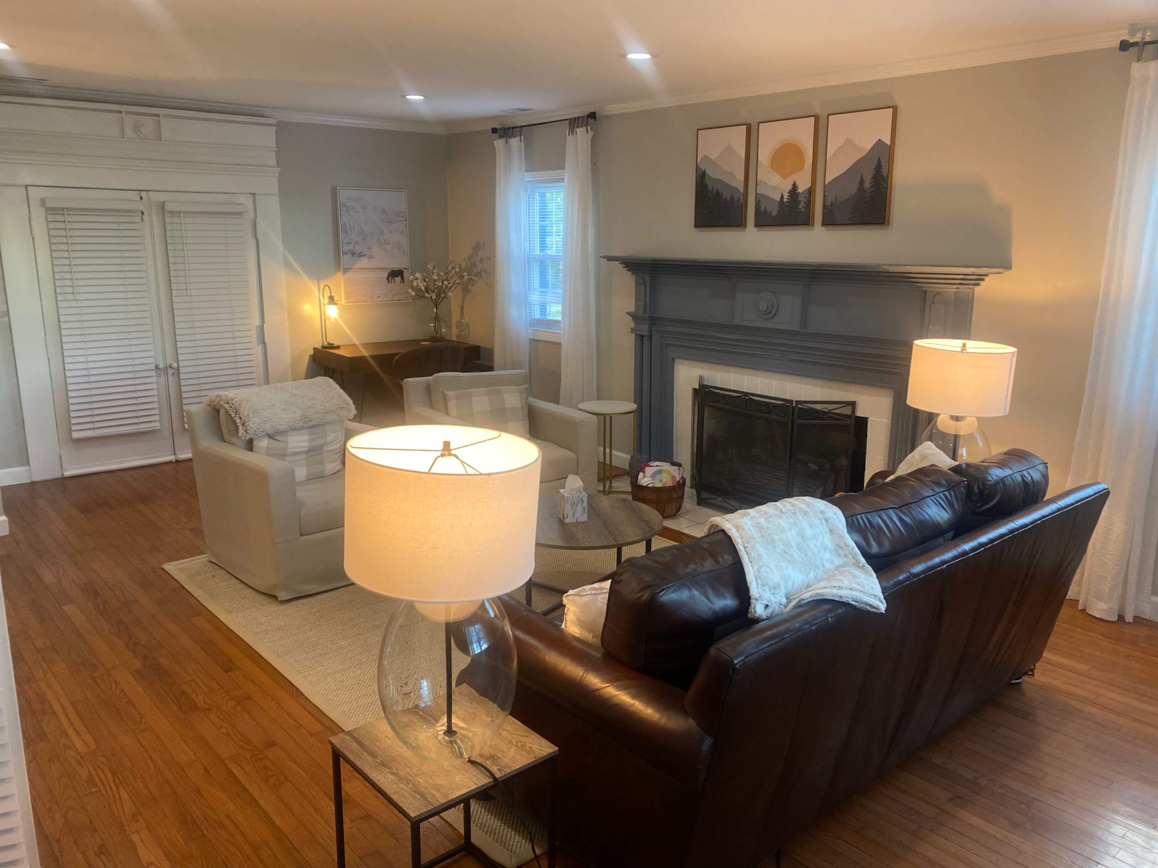 A living room features a brown leather sofa, two light-colored armchairs, a fireplace with a gray mantel, and lamps providing soft lighting.