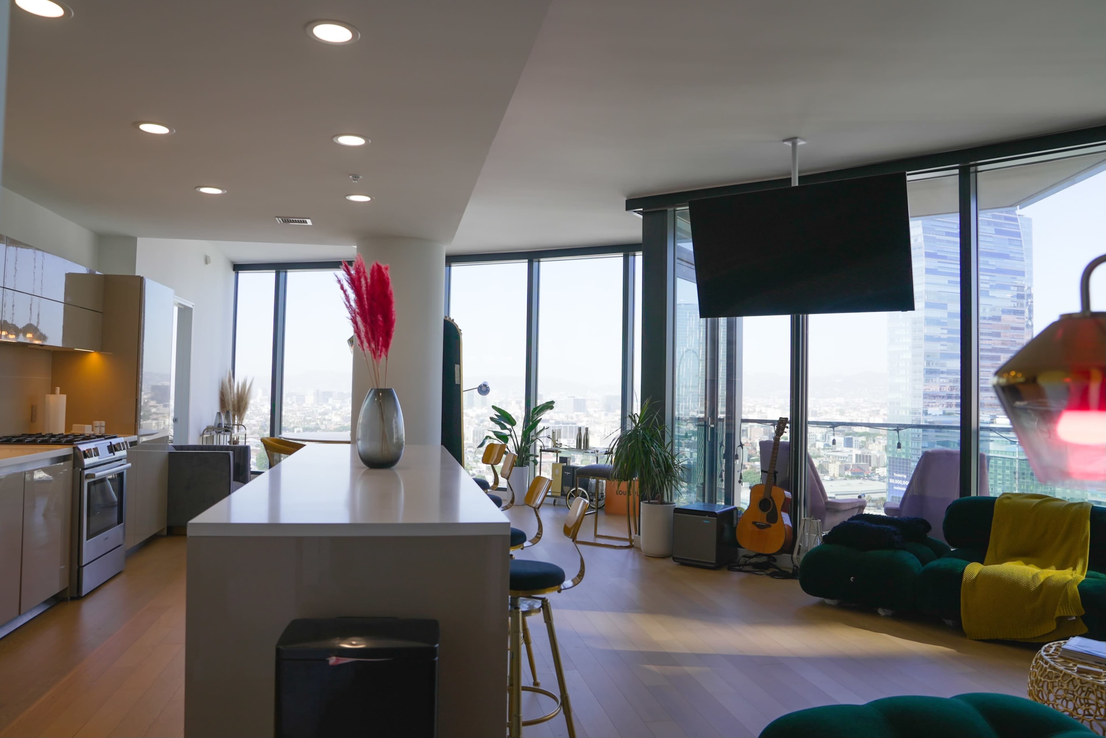 A modern, open-concept apartment features a kitchen with stainless steel appliances, a large island, floor-to-ceiling windows, and a cozy seating area with colorful furniture.