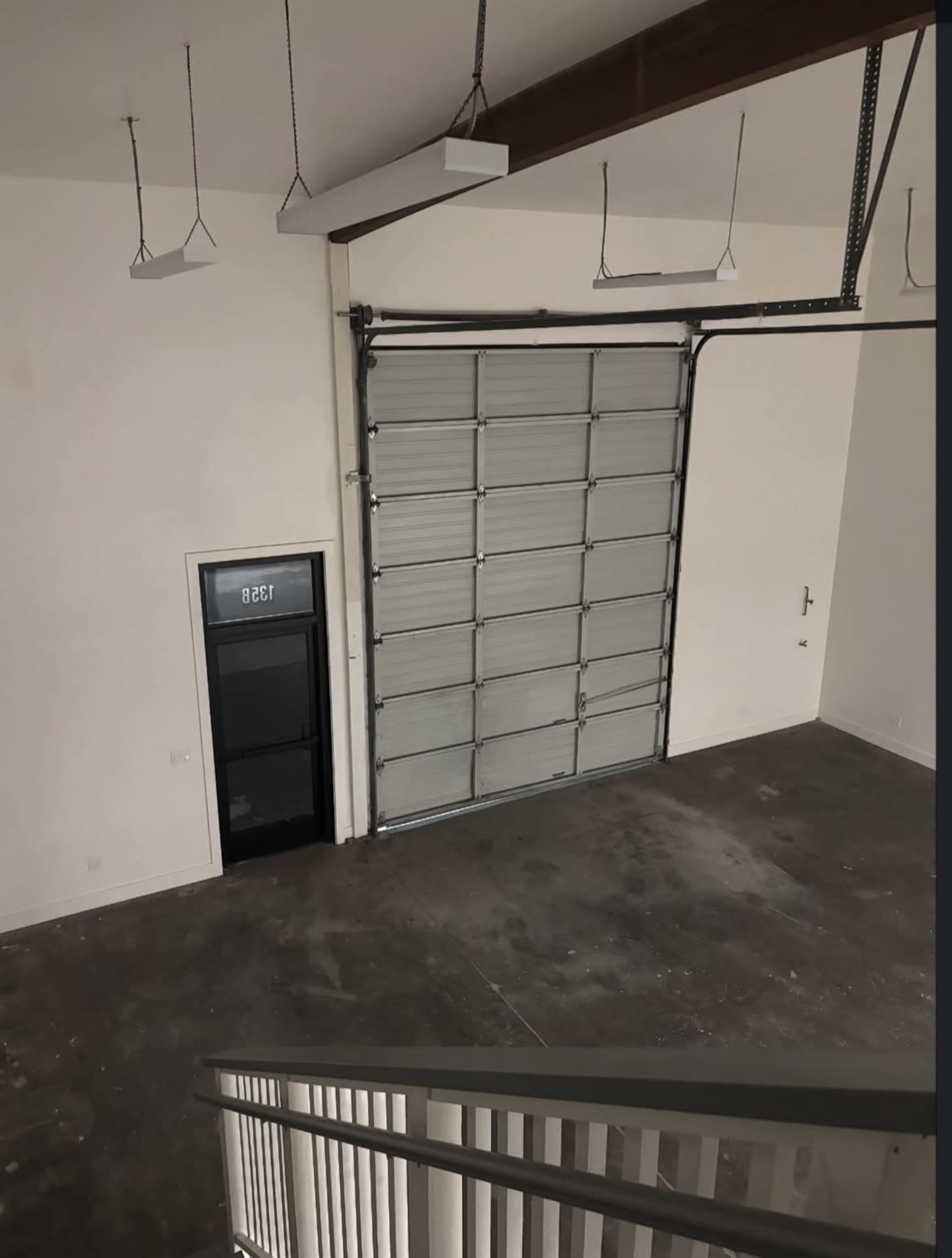 The image shows a spacious and empty garage with a large roll-up door and a glass door on the side, viewed from an upper level.