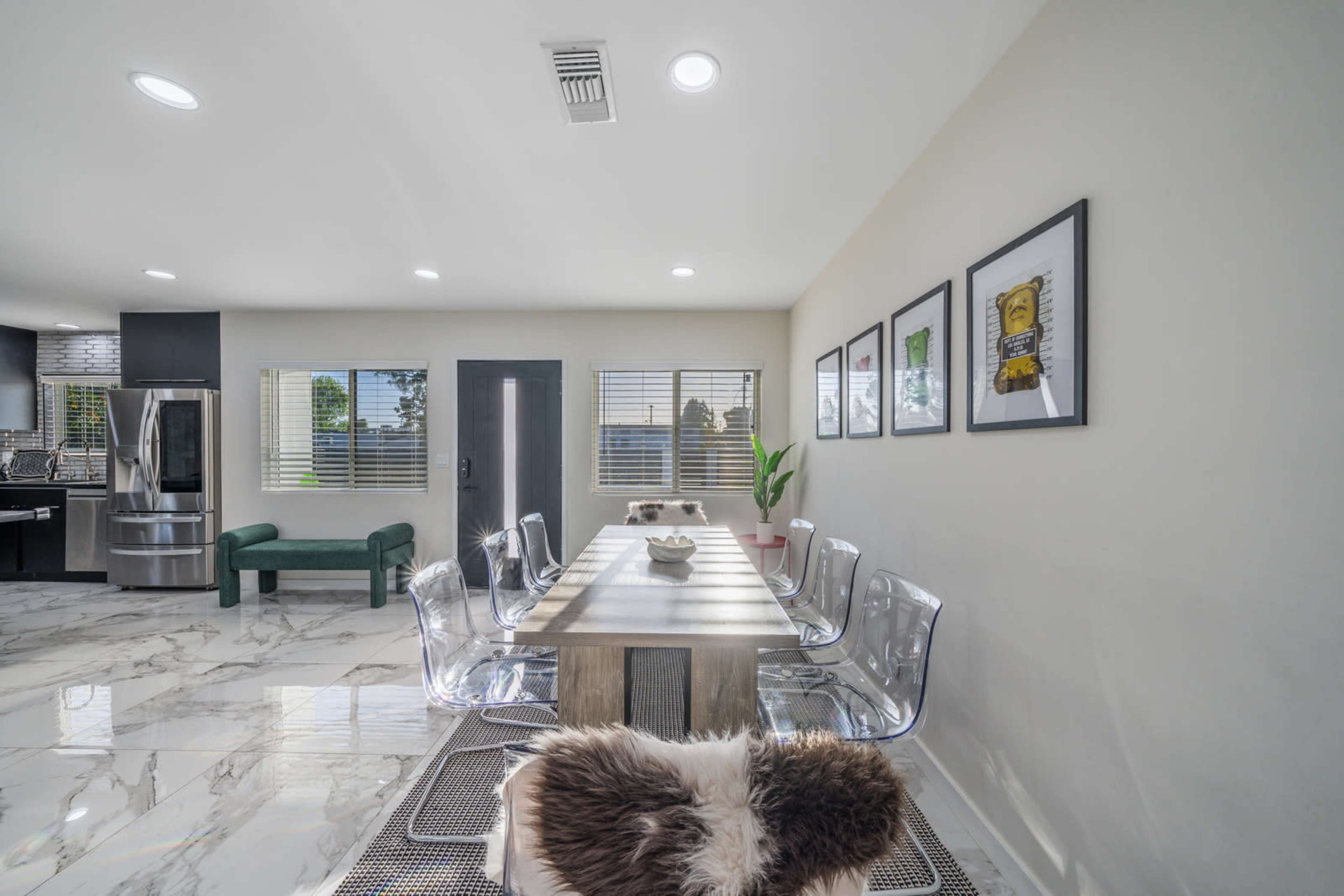 The image shows a modern dining area with a wooden table surrounded by transparent chairs, positioned next to a sleek kitchen and adorned with framed artwork on the wall.