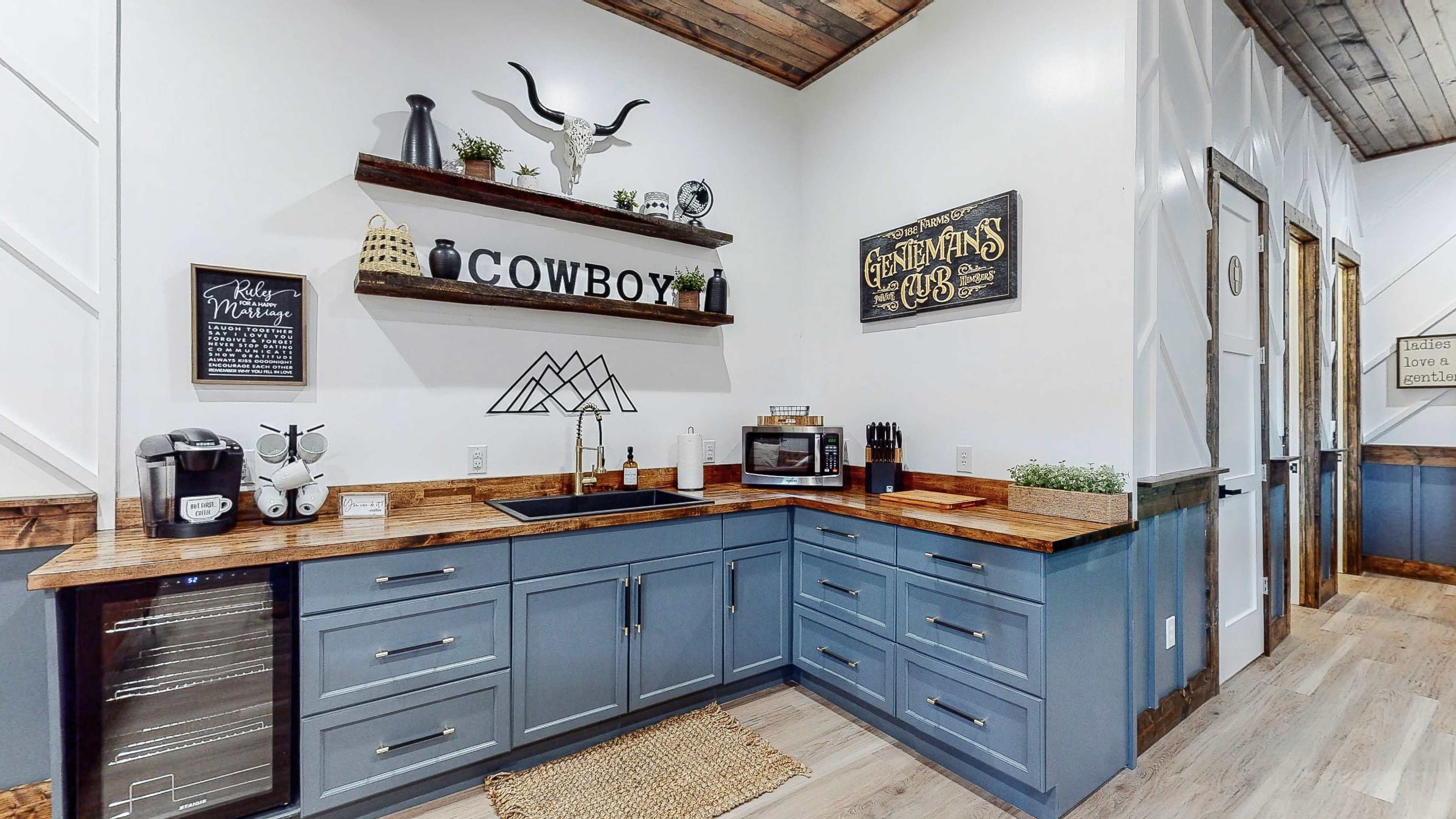 A modern kitchenette with blue cabinetry, a wooden countertop, and wall shelves displaying decorative items and signage.