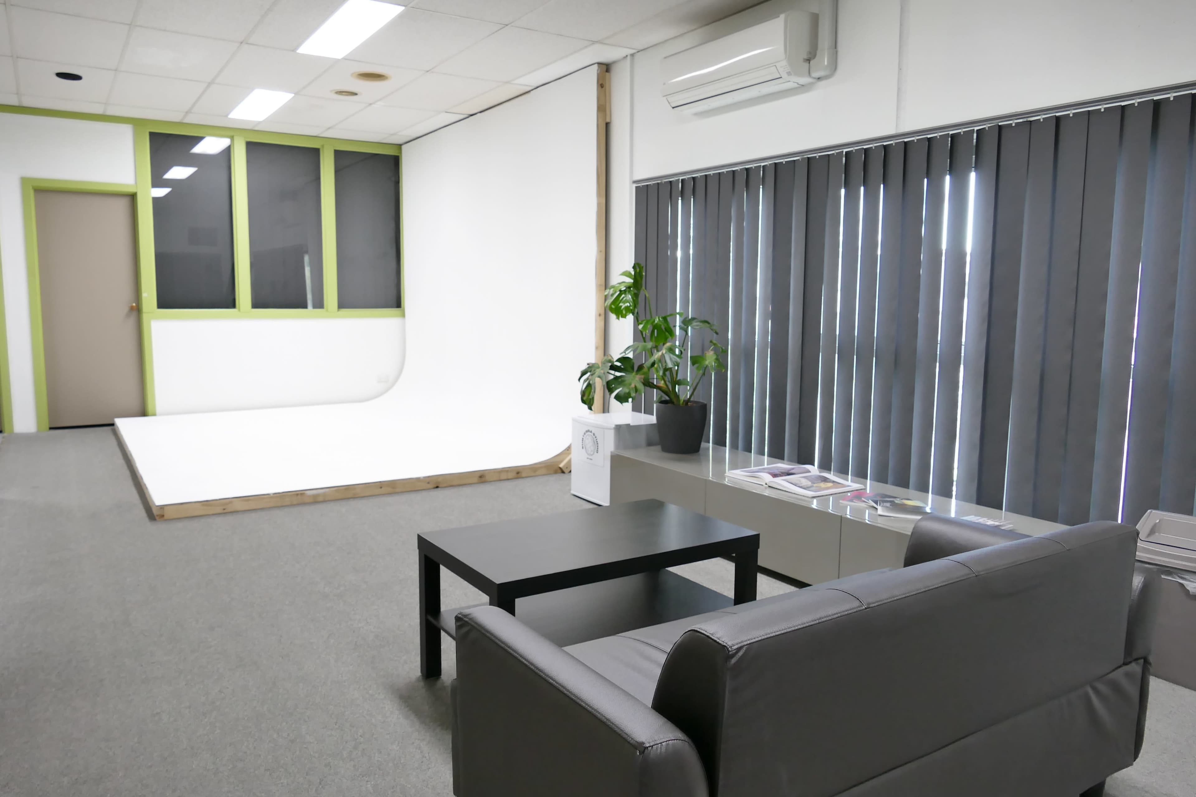 A minimalist studio space with a white backdrop, a black table, a gray couch, and large windows covered by vertical blinds.